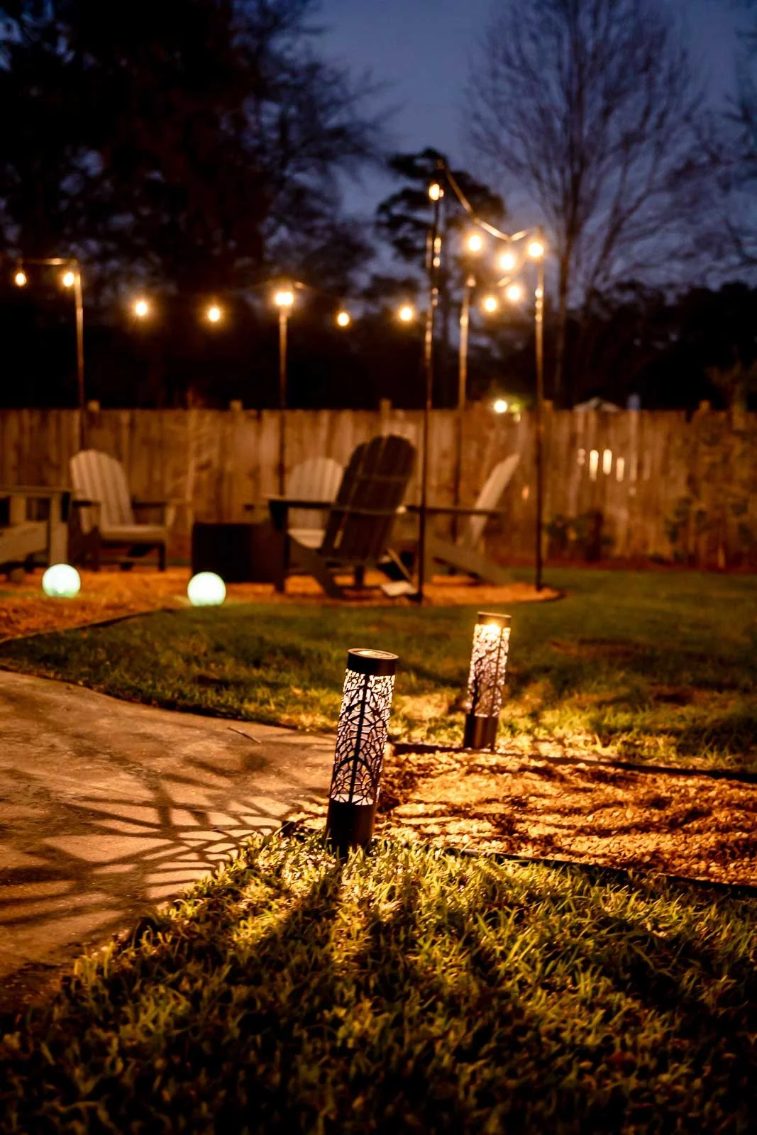 A backyard patio at night illuminated by string lights, with outdoor seating including Adirondack chairs, solar path lights, and glowing globe lights on the ground.