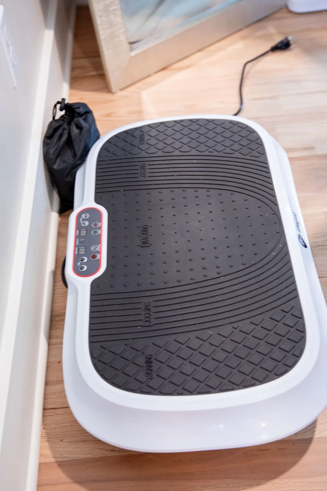 A white and black vibrating platform exercise machine on a wooden floor near a wall with an electrical outlet and a plugged-in power cord.