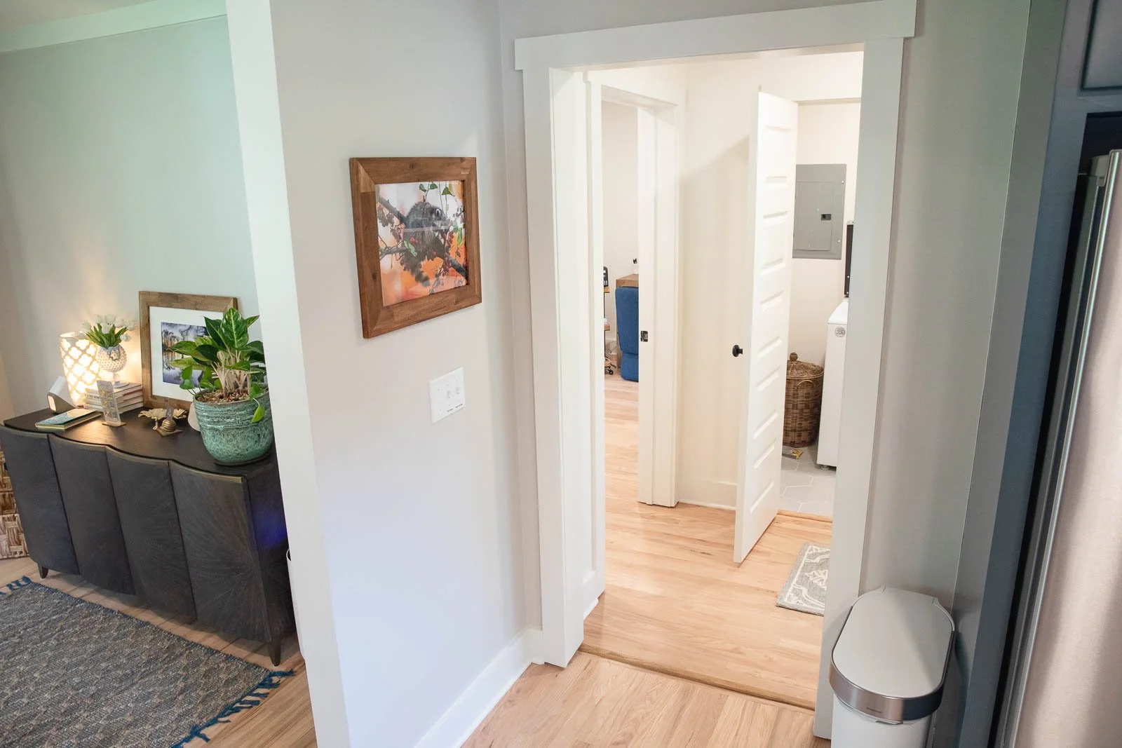 View of a home interior hallway leading to a laundry room with a washing machine and a laundry basket, with a dark cabinet with plants and decorations on the side.