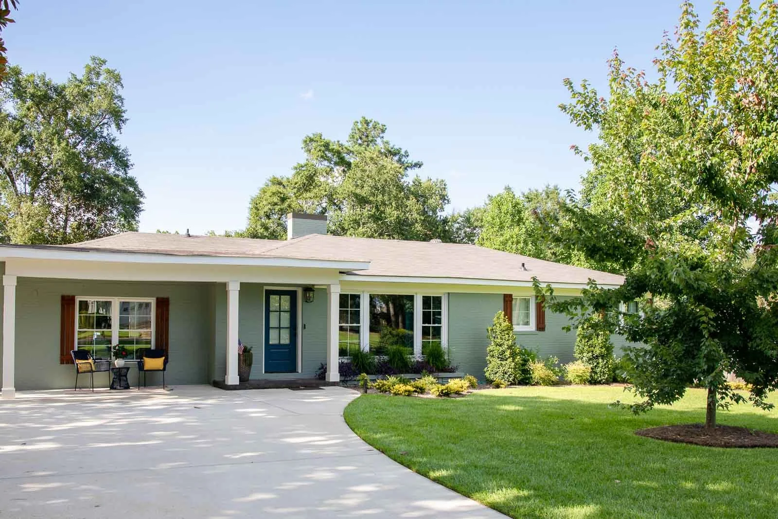 A single-story house with a light green exterior, a front porch, blue front door, and large windows. There is a concrete driveway, green lawn, and trees in the yard.