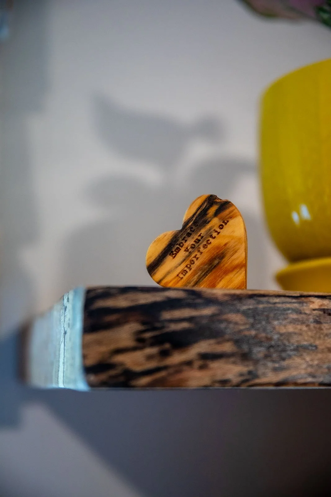 A wooden heart-shaped object with black markings and text, positioned upright next to a piece of wood or bark with a yellow bowl partially visible in the background.