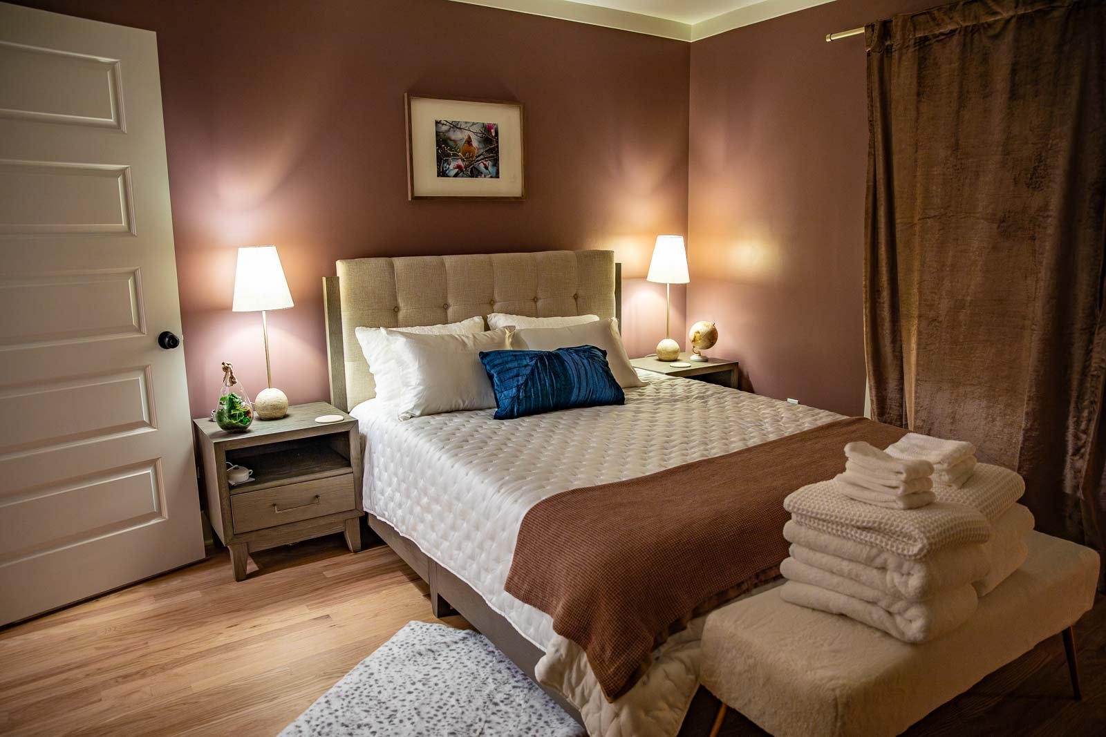 A cozy bedroom with a bed, nightstands on each side, and lamps. The bed is made with white bedding, a blue pillow, and a brown throw blanket. There are folded towels at the foot of the bed. The room has brown walls, a framed picture above the headboa