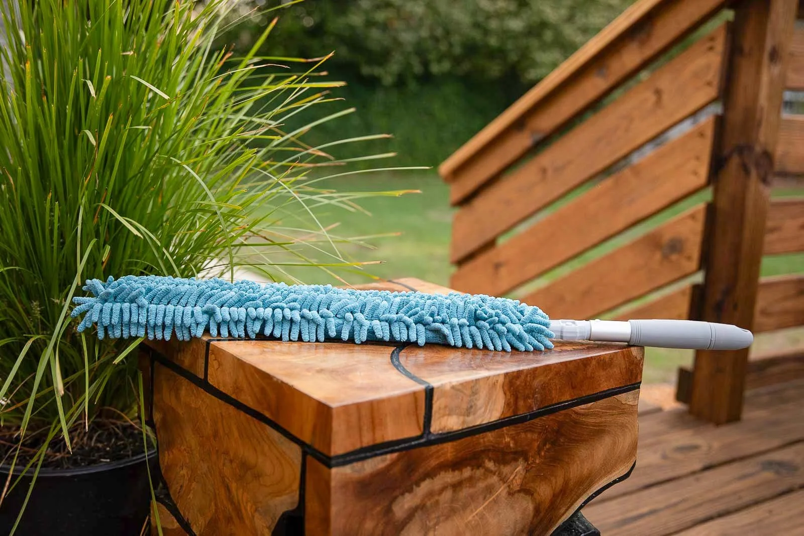 A blue mop resting on a polished wooden bench outdoors, with a green plant and wooden fence in the background.