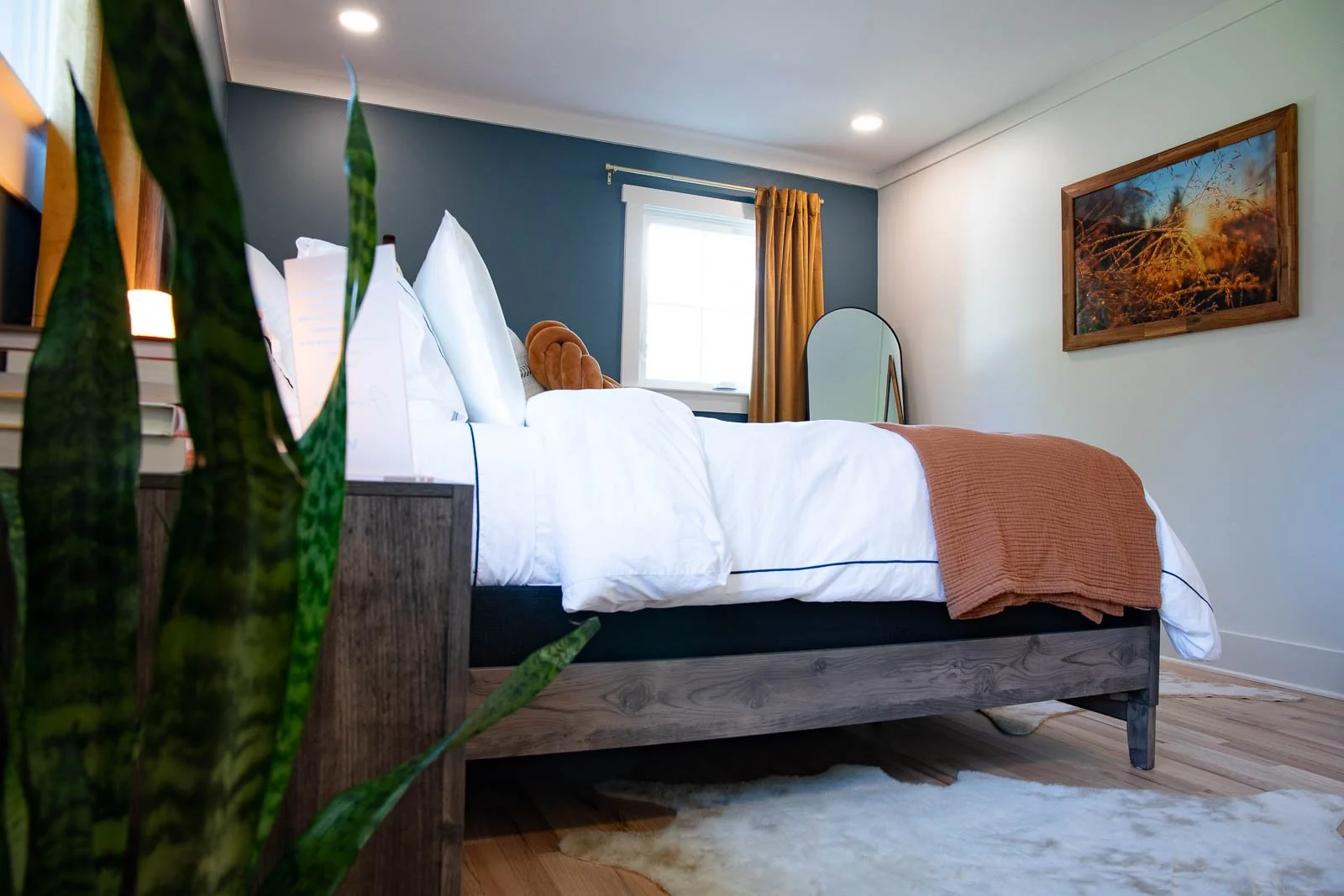 A bedroom with a bed that has white linens and a brown throw blanket, a window with yellow curtains, a wooden picture frame on the white wall, a mirror, and a green plant in the foreground.