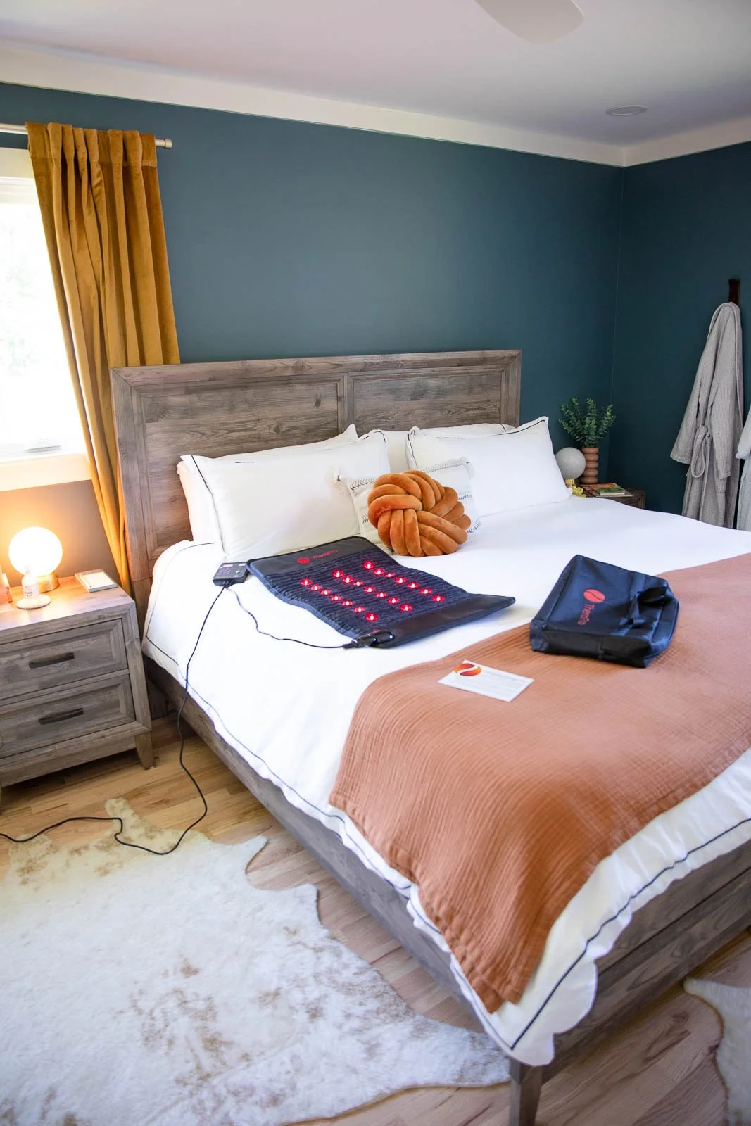 A bedroom with a bed having white pillows, a brown decorative cushion, and a beige blanket. There is a wooden nightstand with a lamp and some items, a teal wall with window curtains, and a robe hanging on the wall. Equipment for a heated blanket or p