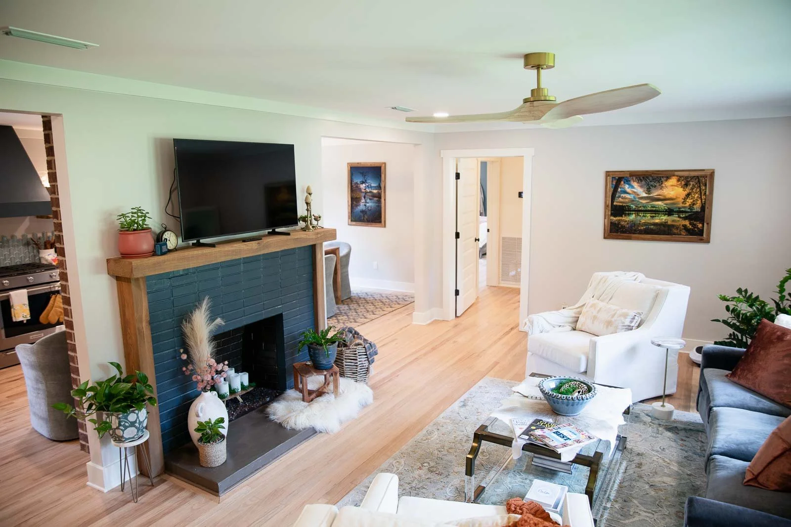 Living room with white walls, hardwood floor, white armchair, dark sofa with pillows, coffee table with magazines and a bowl, artwork on walls, fireplace with blue tiles and plants, ceiling fan, doorway to kitchen area.