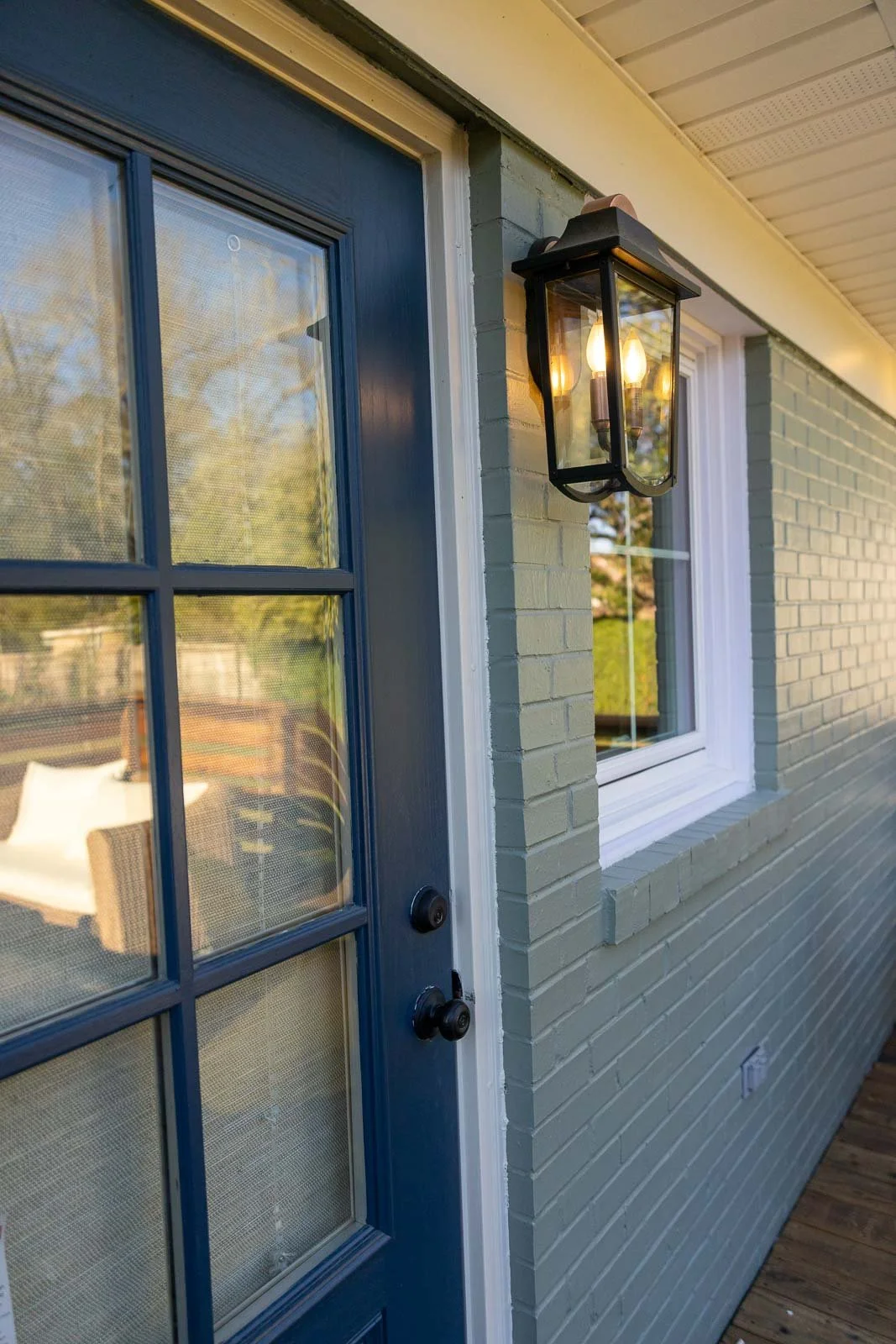 Close-up of a blue door with a window, a black outdoor wall lantern with two bulbs turned on, and a window with a white frame on a light green brick house exterior.