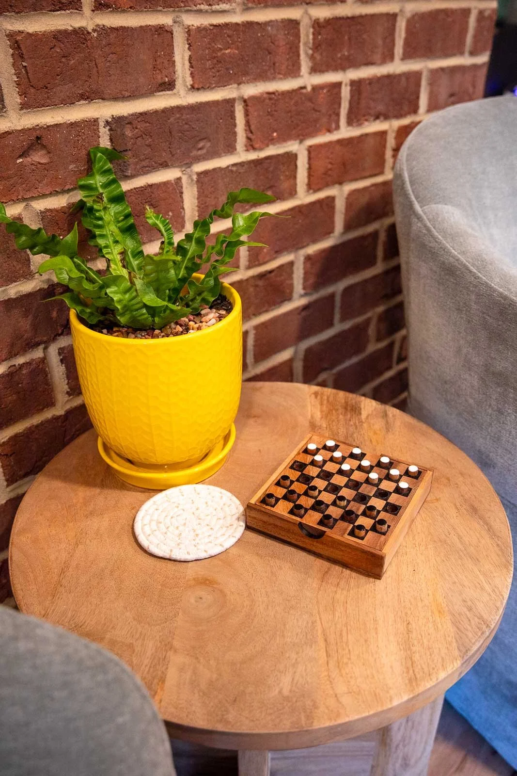 A round wooden side table with a yellow potted plant, a white coaster, and a wooden checkers game set, placed against a brick wall and a gray upholstered chair.