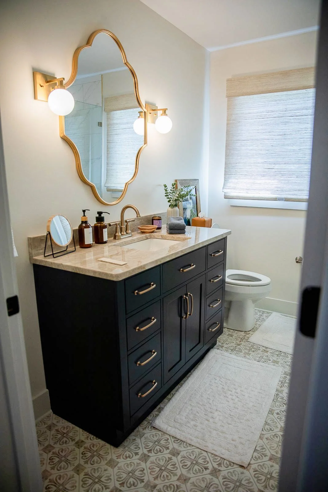Bathroom with a dark vanity, marble countertop, and a mirror with gold frame, two globe light fixtures, a white toilet, two white rugs, decorated with bottles, towels, a plant, and a framed picture, with window blinds.