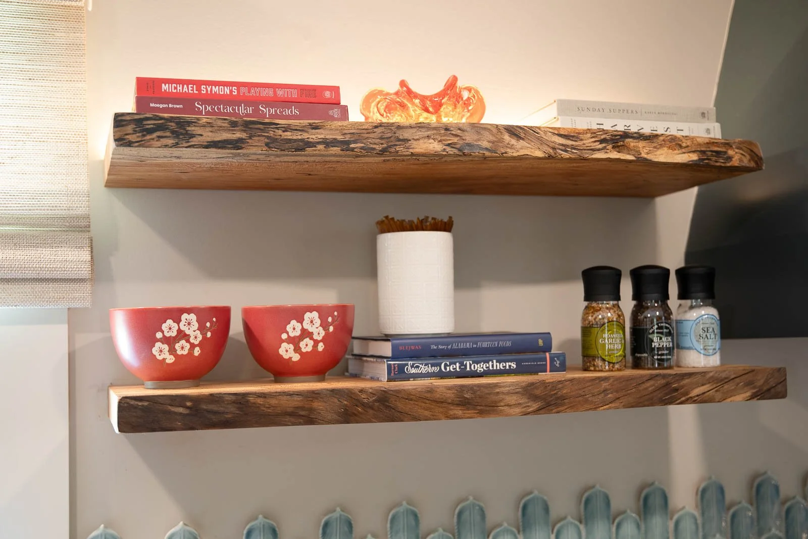 Two wooden shelves on a wall with various items. The top shelf has books titled 'Michael Symon's Playing with Fire', 'Spectacular Spreads', and 'Sunday Suppers', along with a decorative orange and red glass object. The bottom shelf has two pink bowls