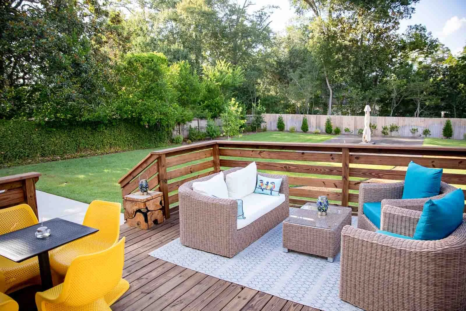 A backyard deck with wicker furniture, colorful cushions, a black dining table with yellow chairs, and a view of a green lawn with trees and a fence in the background.