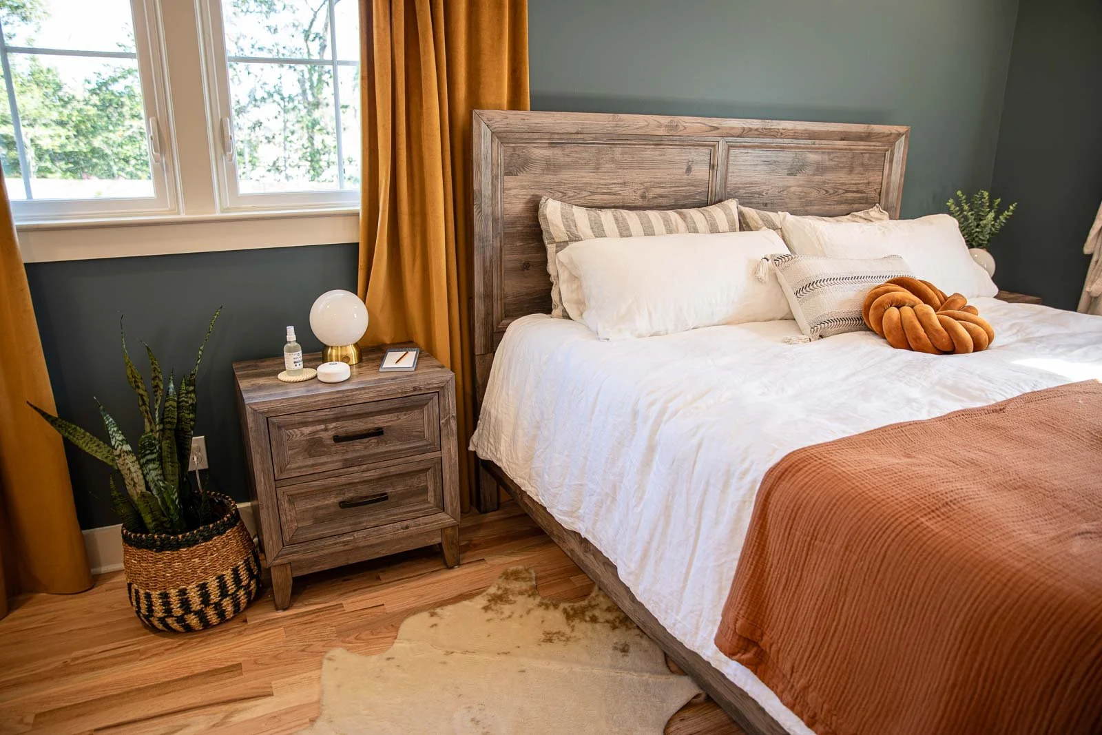 A cozy bedroom with a wooden bed frame, white bedding, and orange accents. A nightstand with a lamp, plants, and small decorative items is beside the bed. There are windows with yellow curtains, hardwood floors, and a rug.