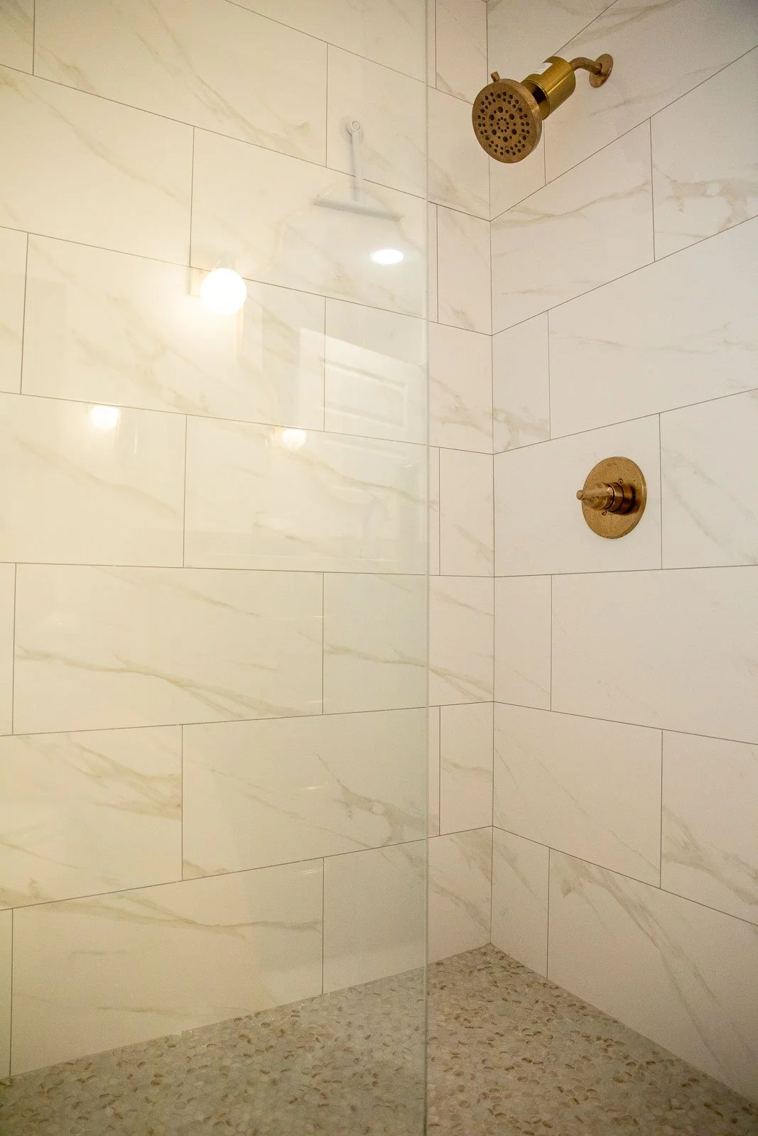 A shower space with beige marble wall tiles, a bronze showerhead, and a bronze control handle, separated by a glass panel on the left side.