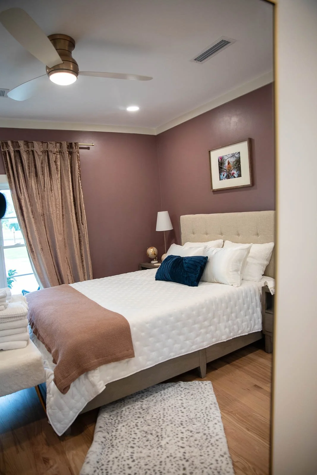 A neatly made bed with white bedding, a beige tufted headboard, and a navy pillow, in a bedroom with burgundy walls, framed artwork, a nightstand with a lamp, and a window with gold curtains.