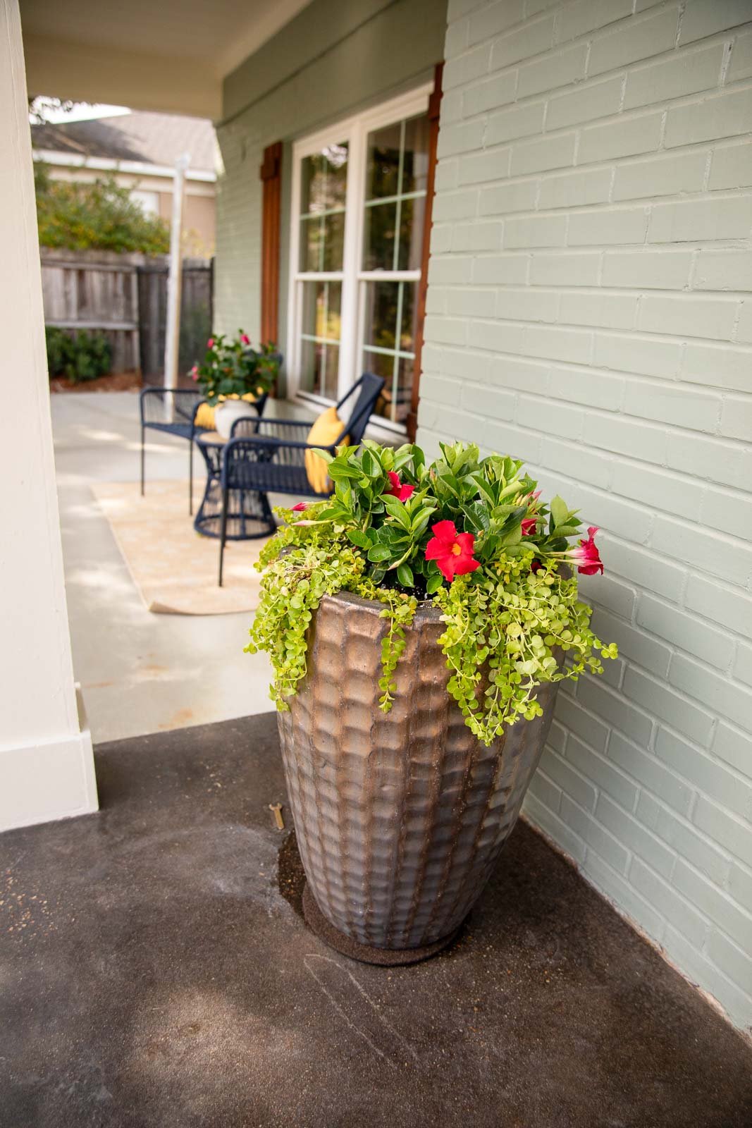 Large textured pot with red and pink flowers and green foliage on a porch with outdoor seating visible in background.