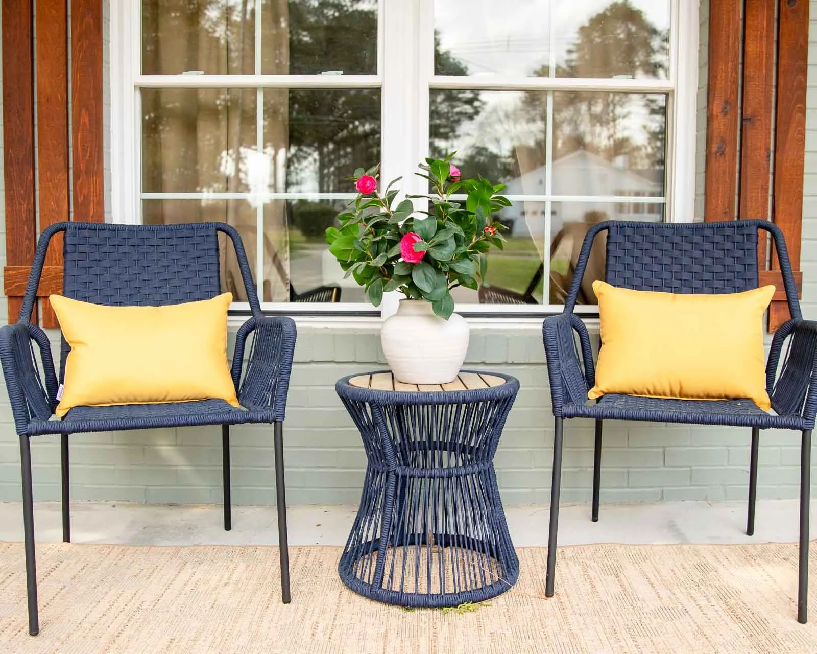 Outdoor patio with two navy blue wire chairs with yellow pillows, a navy blue round side table, and a white pot with pink flowers, in front of a house window with wooden trim.