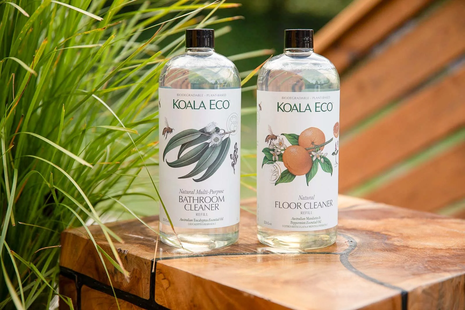 Two bottles of Koala Eco cleaning products, a bathroom cleaner and a floor cleaner, placed on a wooden surface outdoors with green plants in the background.