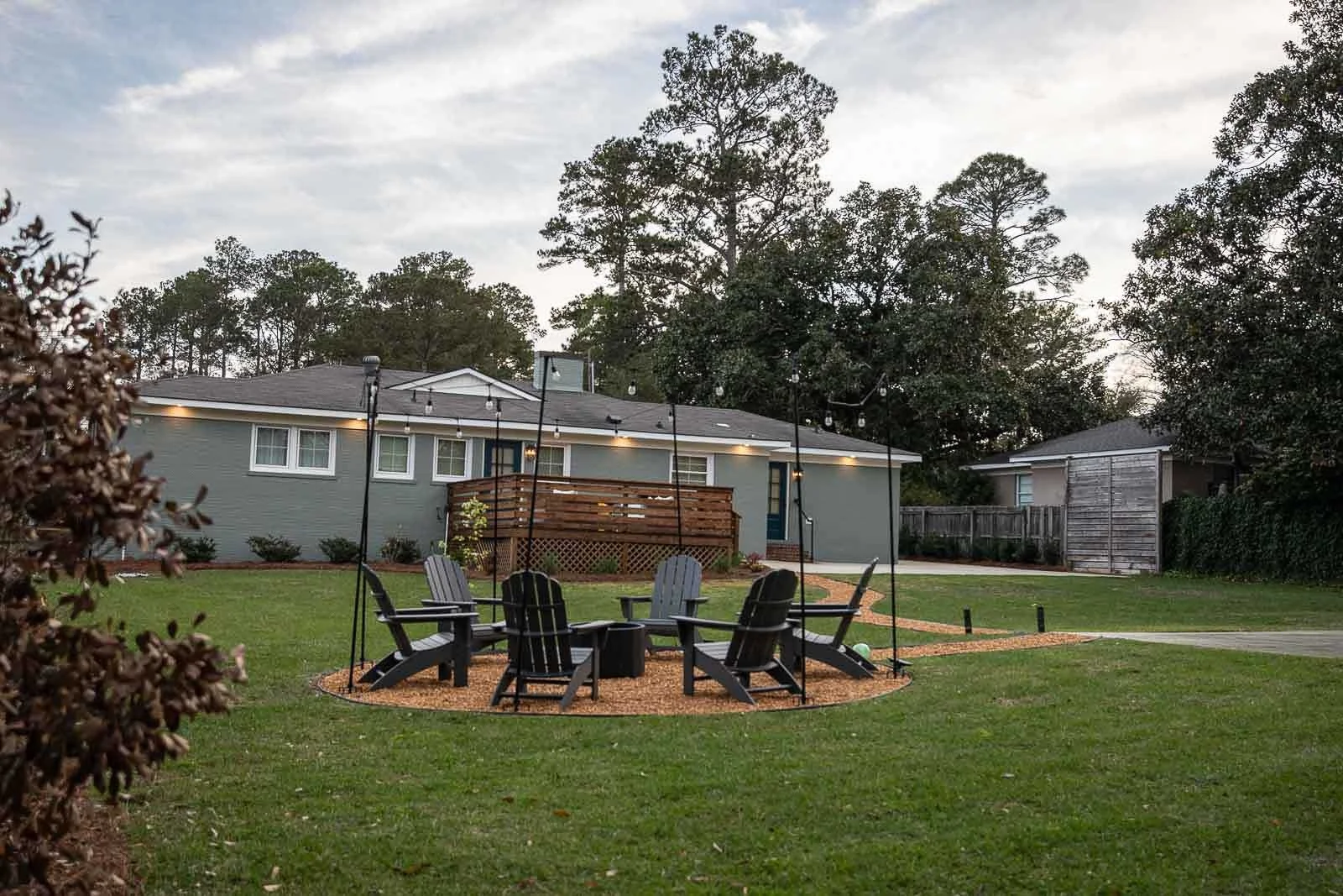 A backyard with a circle of six black Adirondack chairs surrounding a fire pit on a gravel surface. There is a wooden deck attached to the gray house, string lights hanging overhead, and trees in the background on a cloudy day.