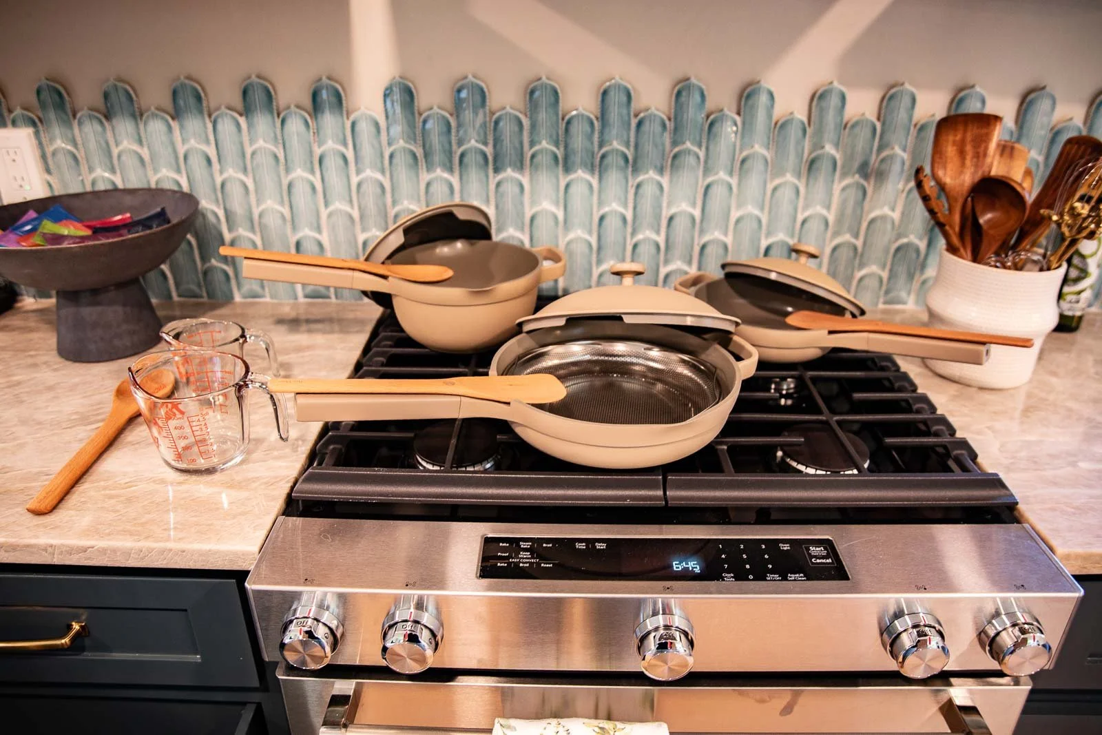 A stove with multiple stacked oven-safe pots and pans, some with wooden utensils, a measuring cup, and a wooden spoon nearby. A decorative tile backsplash and a container with cooking utensils are in the background.