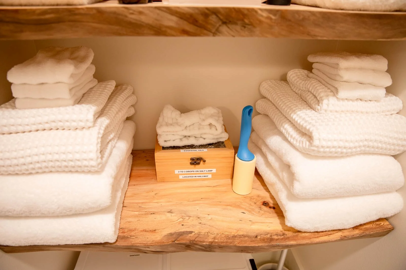 Stacks of white towels on wooden shelves in a linen closet or bathroom cabinet, with a lint roller and a small box labeled for essential oils.
