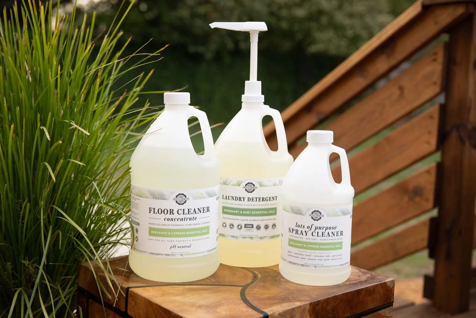Three bottles of cleaning products on a wooden table outdoors, with greenery and a wooden railing in the background. The bottles are labeled for floor cleaner, laundry detergent, and spray cleaner.