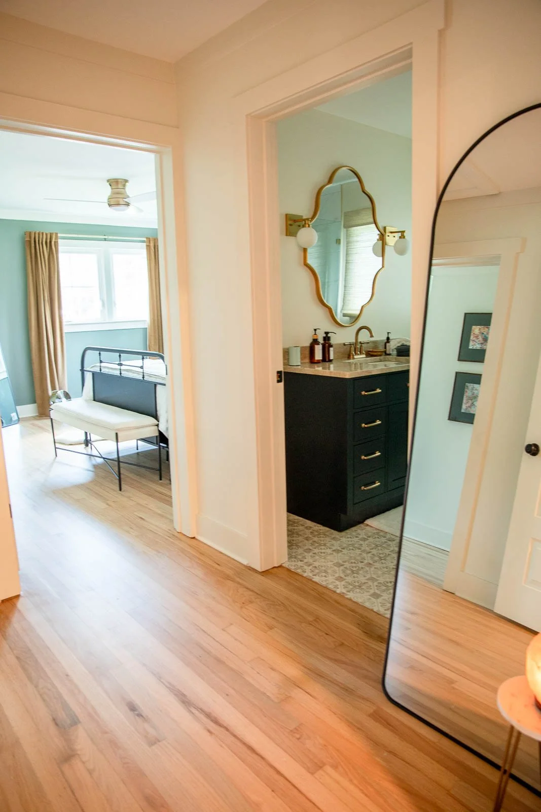 View of a hallway with a bathroom and bedroom visible. The bathroom has a black vanity with gold handles, a gold-framed mirror, and bottles on the sink. The bedroom has a metal bed frame, a bench, and a window with yellow curtains.