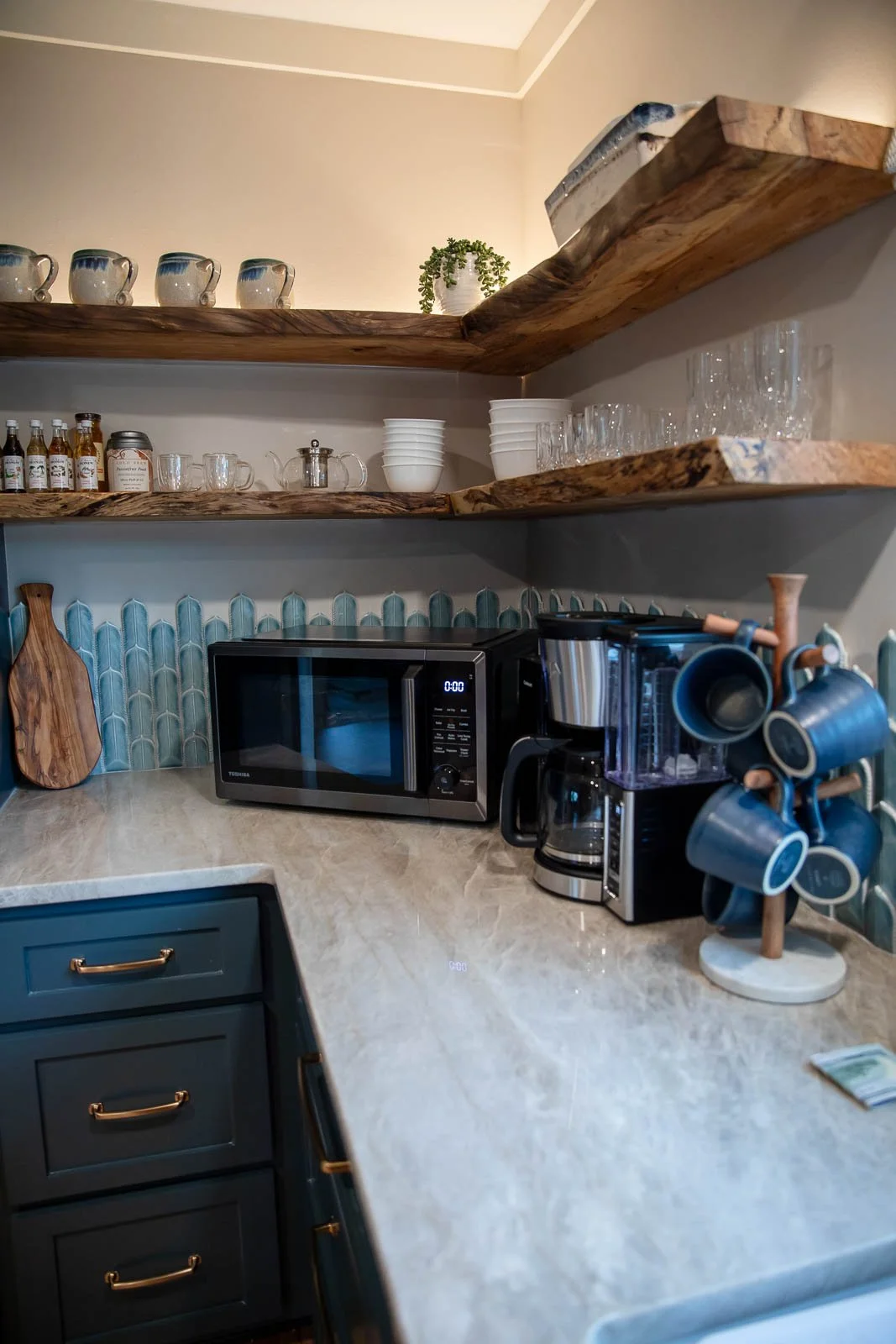 Kitchen counter with a microwave, coffee maker, mugs, wooden cutting board, and open shelves with cups, glasses, and small bottles.