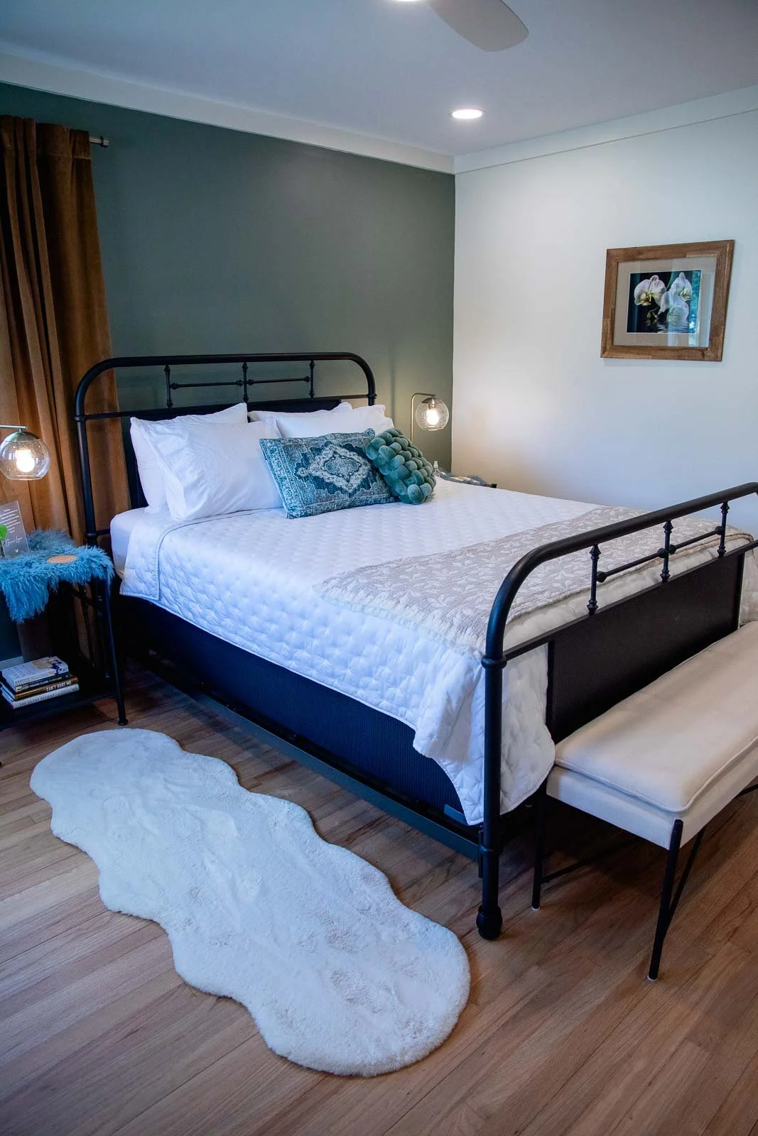 A bedroom with a black metal bed frame, white bedding, decorative pillows, a white furry rug, and wooden flooring. There are two hanging bedside lamps and a small side table with books and a blue furry cover. The wall behind the bed has a two-tone pa