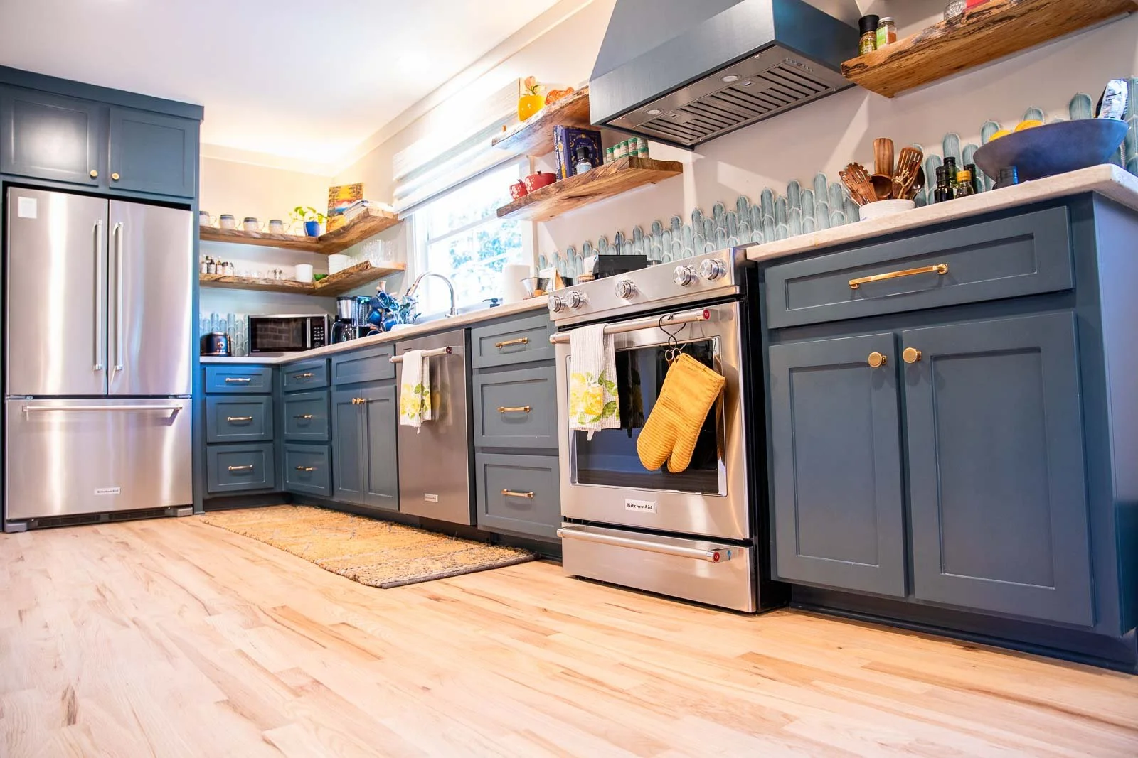 Modern kitchen with blue cabinets, stainless steel appliances, open wooden shelves, and a window above the sink.