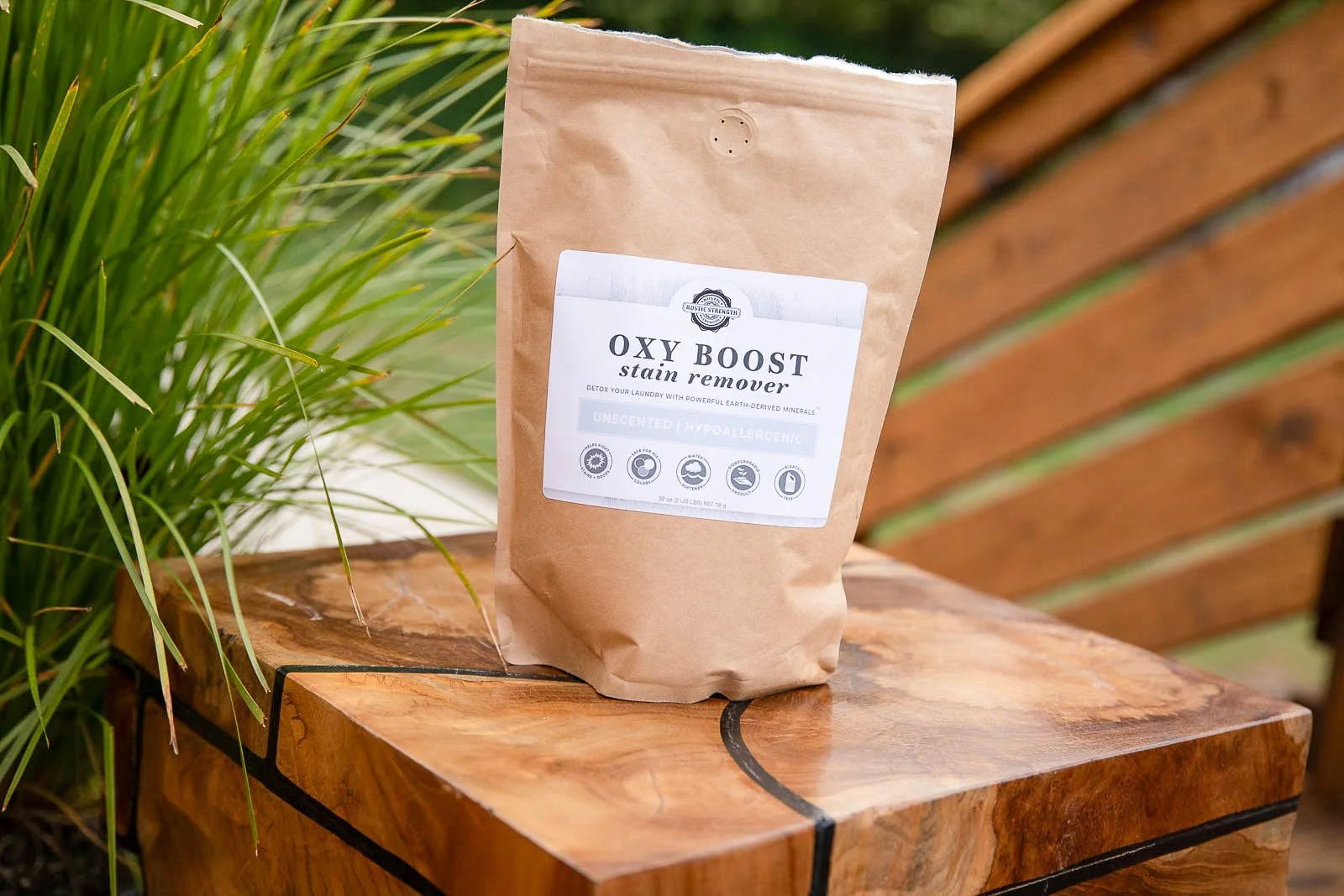 A brown paper bag of Oxy Boost stain remover on a wooden table outdoors, with green plants and a wooden fence in the background.