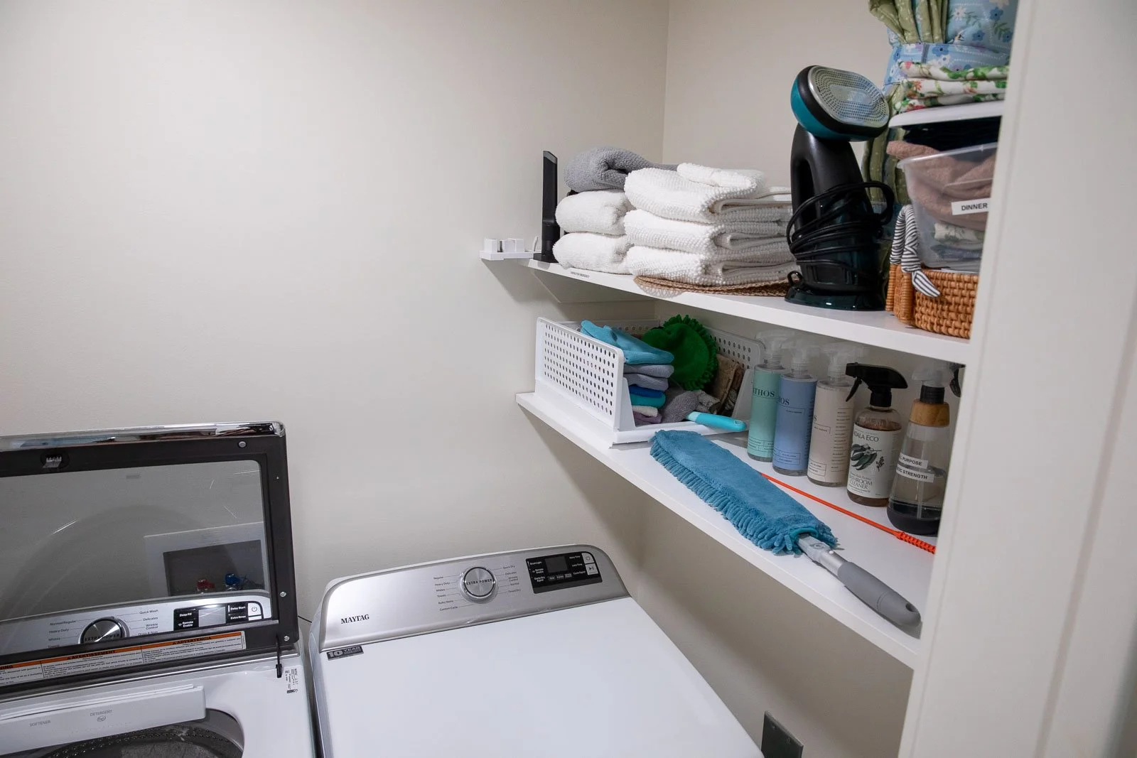 Laundry room with shelves holding towels, cleaning supplies, and miscellaneous items, next to a washing machine.