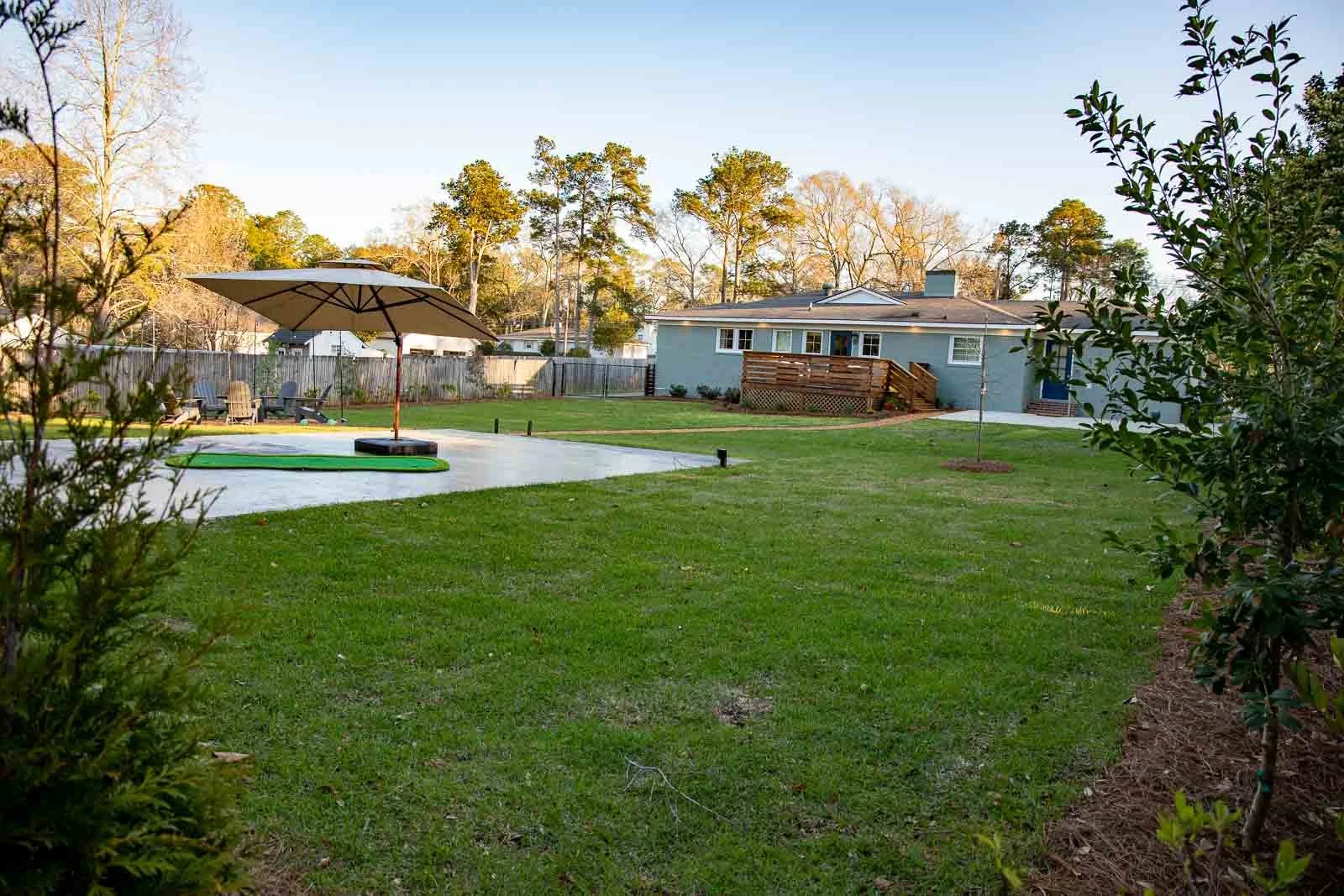 A backyard with a grassy lawn, patio area with an umbrella, outdoor chairs, and a house in the background. There are trees and a wooden fence surrounding the yard.