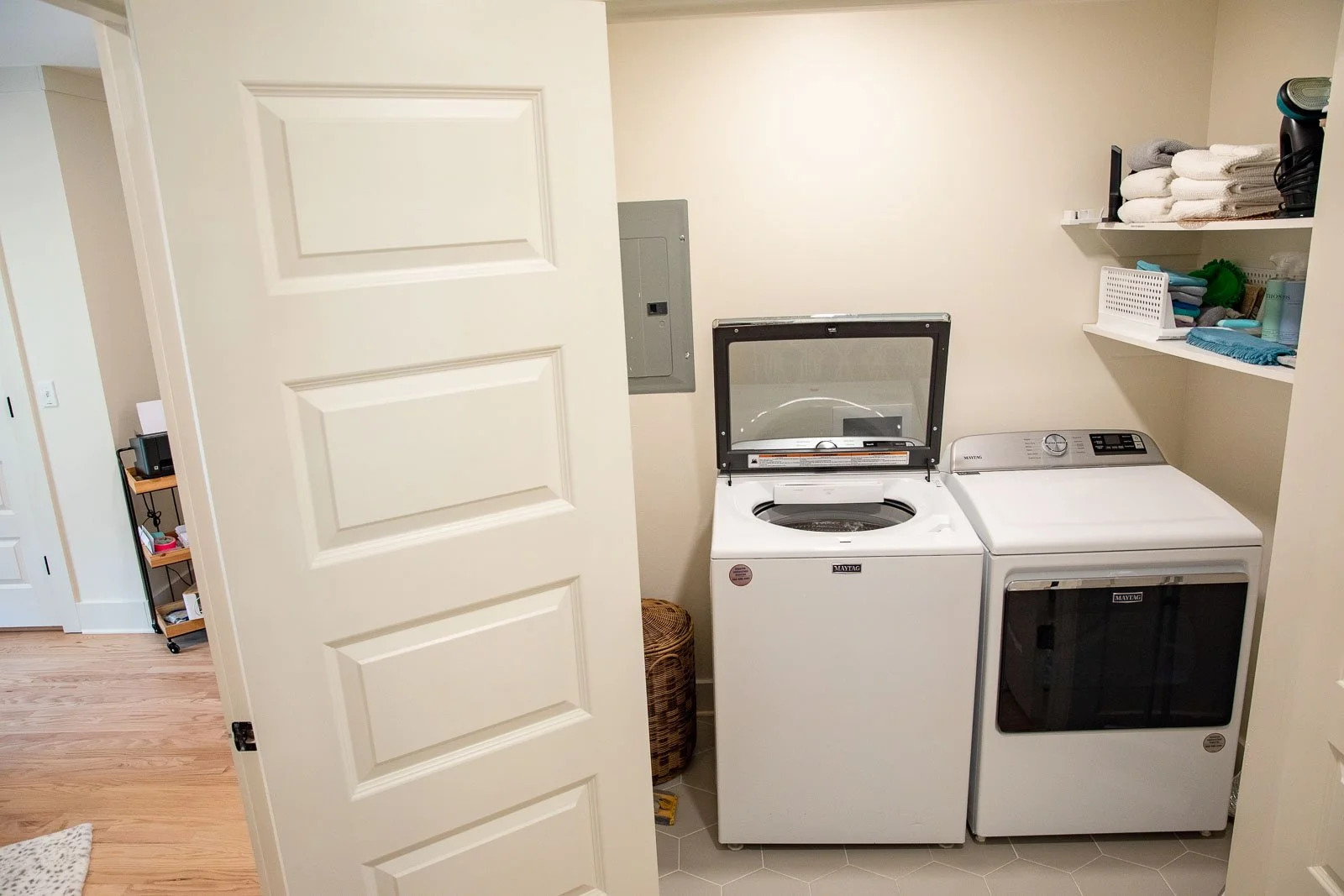 Laundry room with a top-loading washing machine and a front-loading dryer side by side, shelves with folded towels and laundry supplies above them.