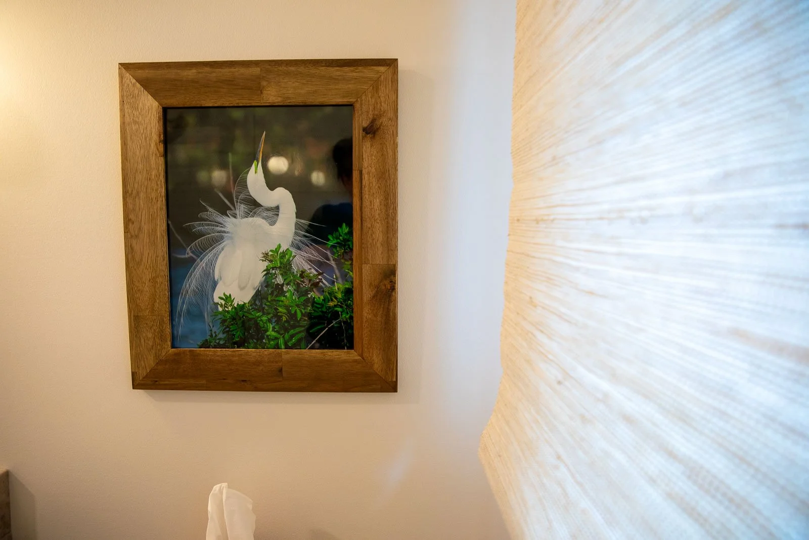 A framed picture of a white heron standing among green foliage, with a wooden frame, hung on a beige wall next to a light-colored textured curtain or wall covering.