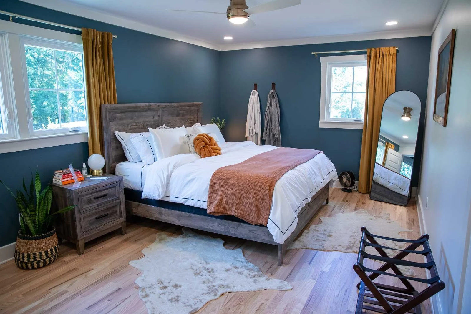 A cozy bedroom with a wooden bed frame, white bedding, and an orange throw blanket. Two windows with yellow curtains, a nightstand with books, a lamp, and a plant. A full-length mirror leans against the wall, and two bathrobes hang on hooks. A small 