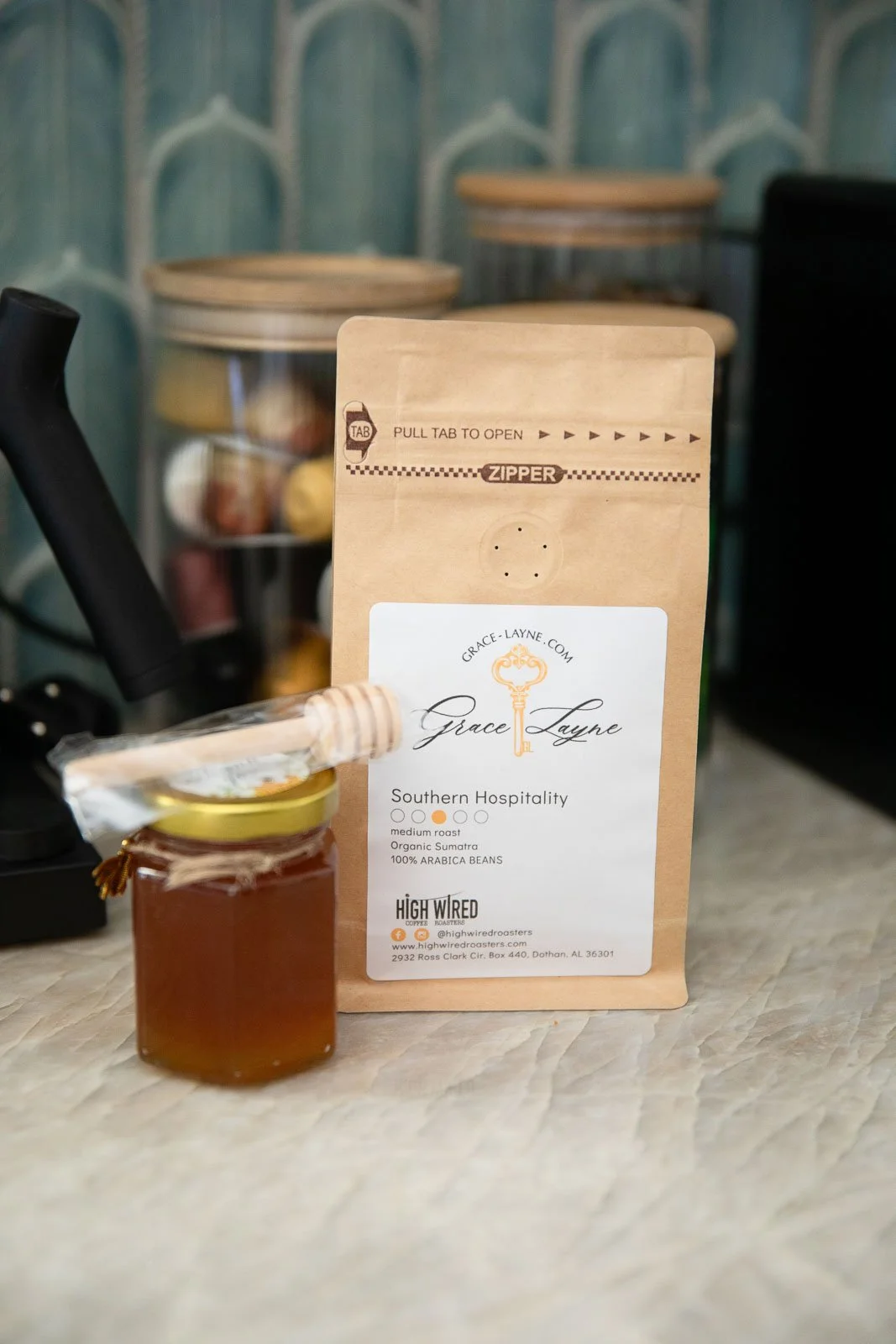 A small jar of honey with a honey dipper on top, in front of a package of Grace Layne coffee beans, with coffee canisters in the background.