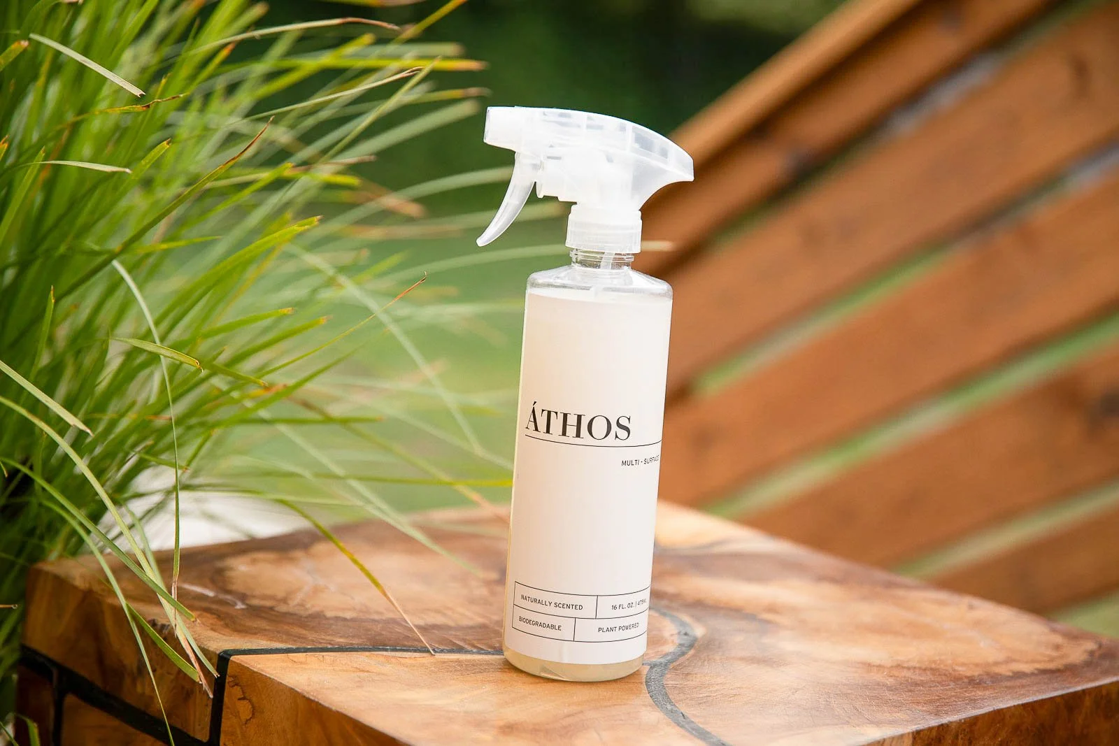 A spray bottle labeled 'ÁTHOS,' placed on a wooden surface outdoors, with green plants and a wooden bench in the background.