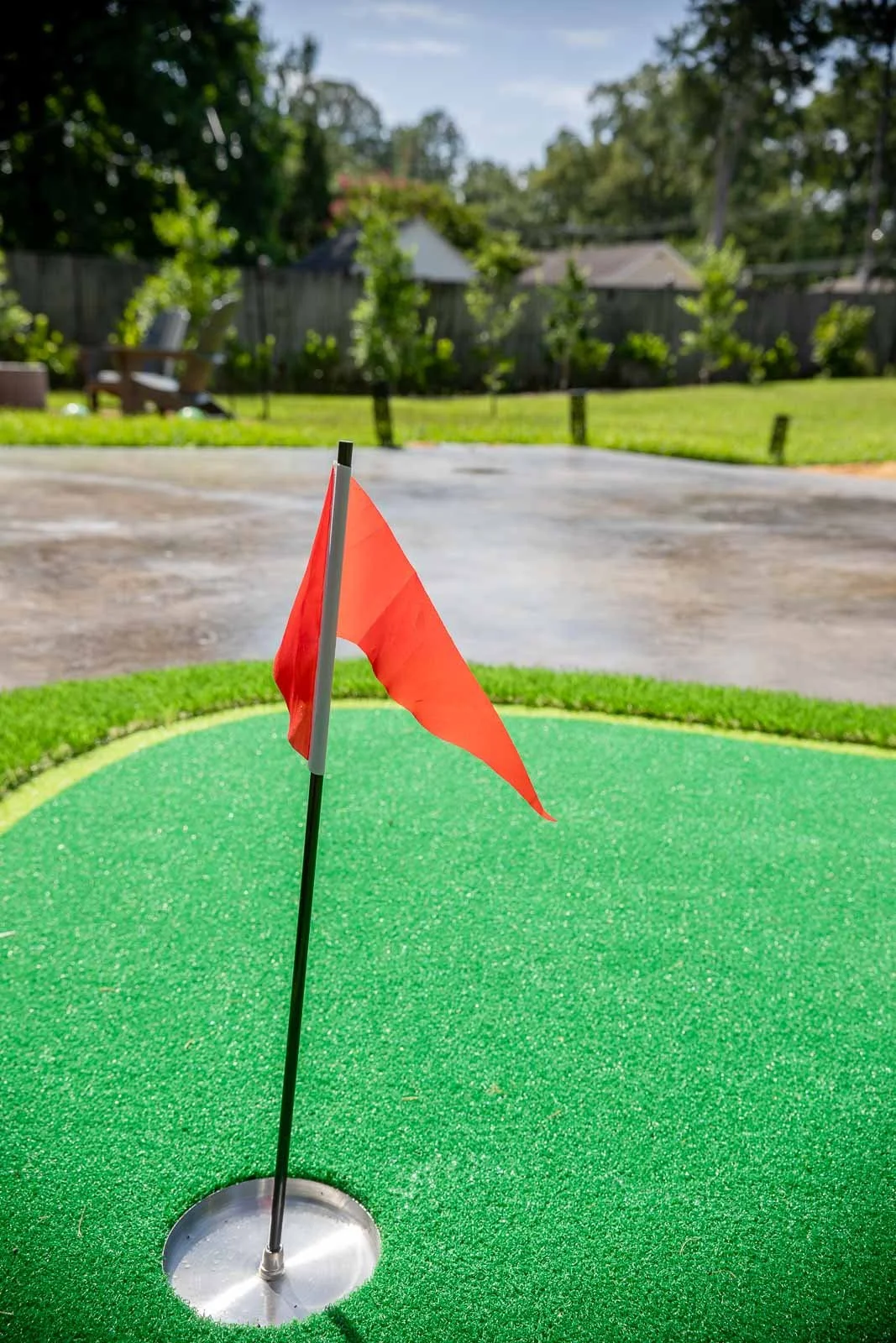 Miniature golf hole with a red flag on a green artificial turf, with outdoor backyard in the background.