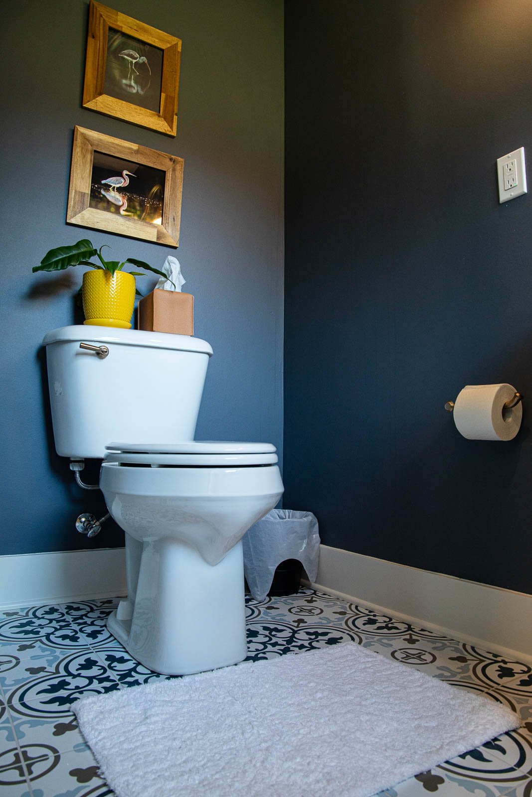A bathroom with a white toilet, a white rug, a trash bin, a toilet paper holder, and two framed pictures of flamingos on the dark gray wall. There is a yellow plant pot with a green plant and a tissue box on top of the toilet tank.