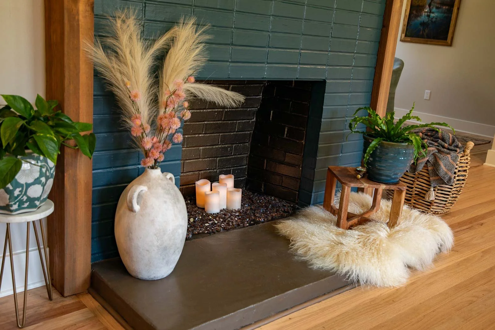 Living room with a dark teal fireplace, white candles inside, a large white vase with pink and beige pampas grass on the left, a wooden side table with a potted green plant on the right, a fluffy cream-colored rug underneath, and a wicker basket with