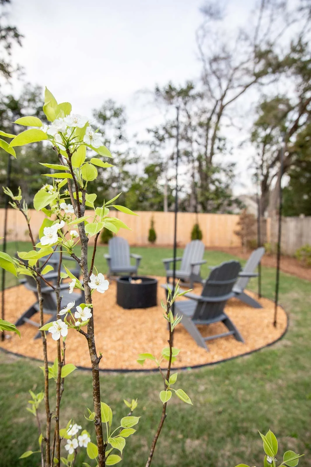 A backyard with a circular gravel fire pit surrounded by wooden Adirondack chairs, enclosed by a wooden fence, with trees and a cloudy sky in the background.