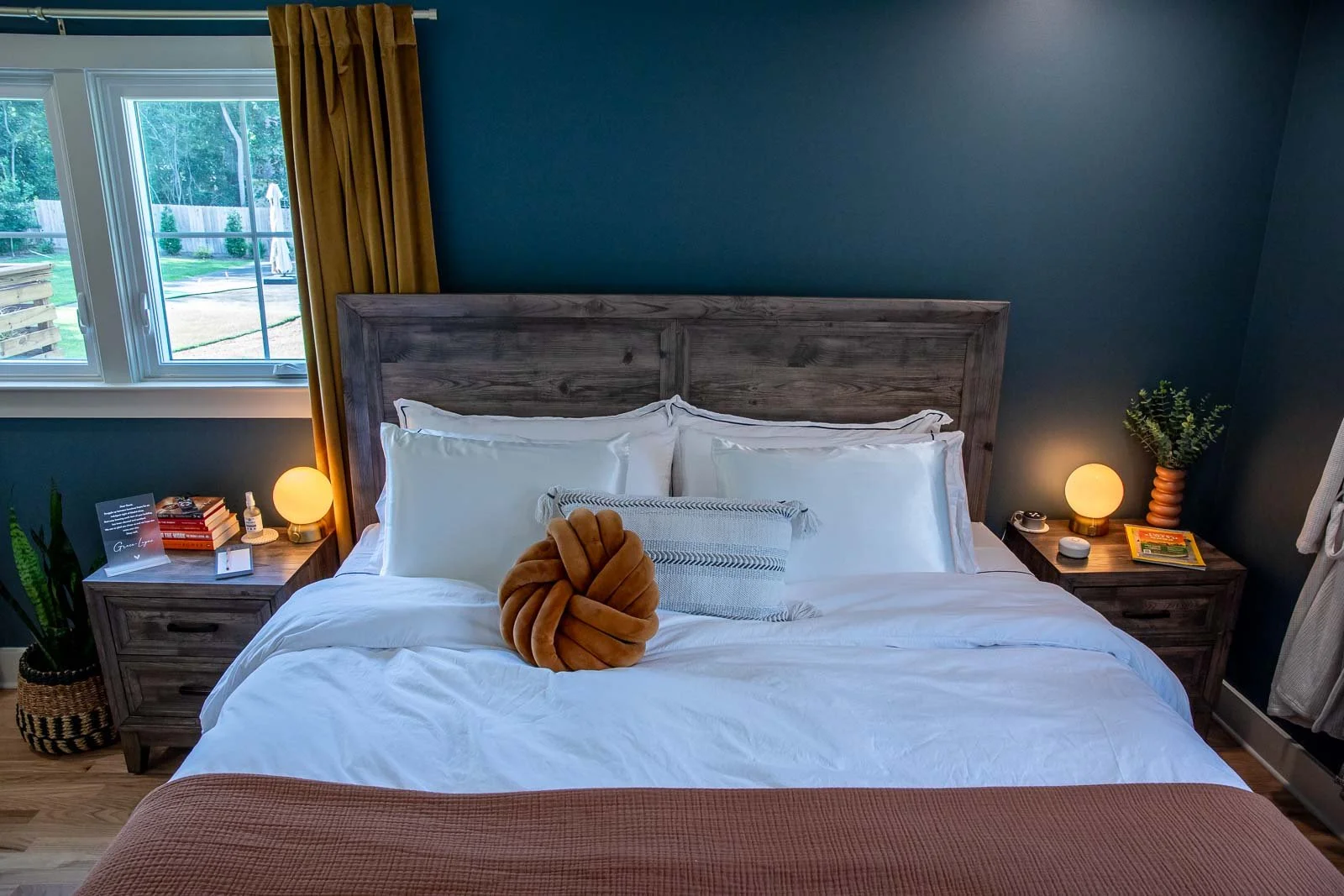 A cozy bedroom with a large bed with white bedding and a tan knot pillow, wooden headboard, and matching nightstands. There are small lamps, plants, books, and a window with gold curtains showing a backyard.