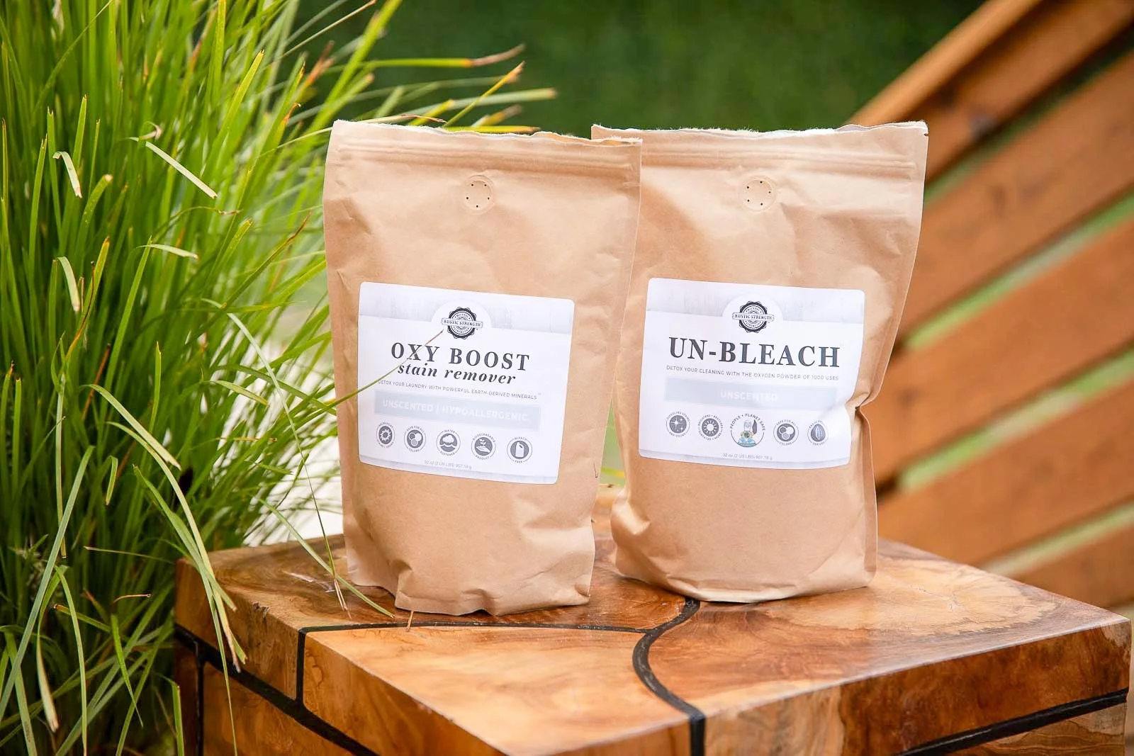 Two brown paper bags of cleaning products labeled 'OXY BOOST stain remover' and 'UN-BLEACH' sit on a wooden surface outdoors next to green plants and a wooden fence.