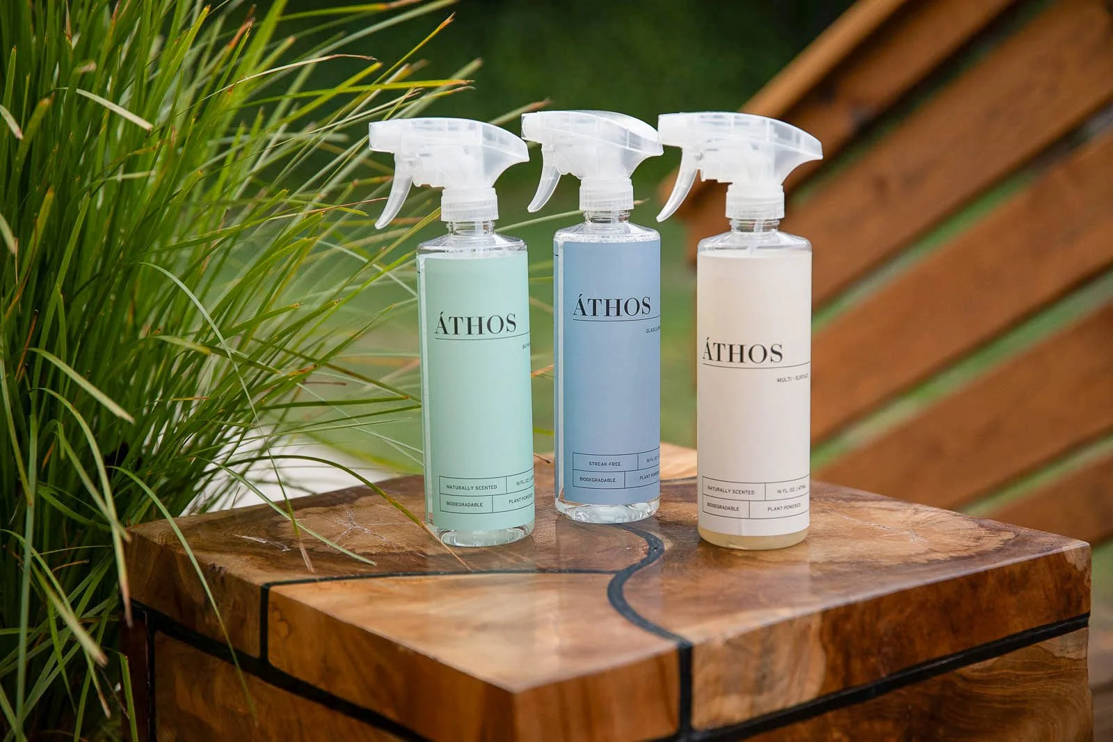 Three spray bottles labeled 'Athos' on a wooden surface, with green plants on the left and a wooden background on the right.