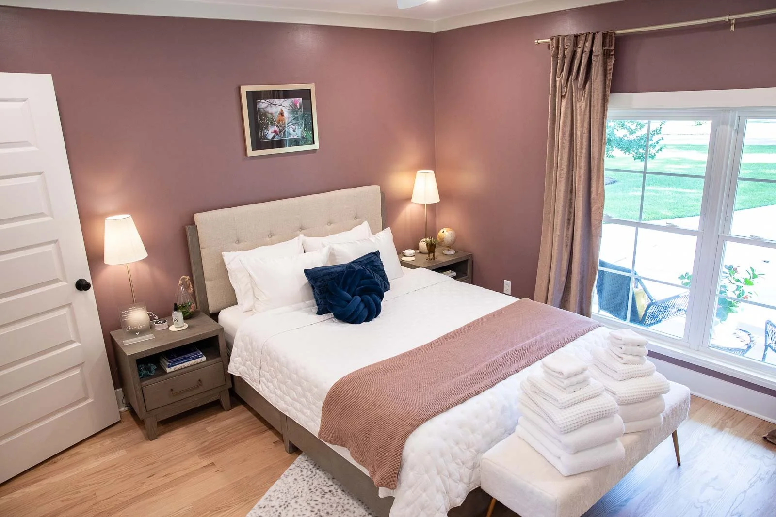 A cozy bedroom with a large bed dressed in white bedding, a navy pillow, and a brown throw blanket. Two matching nightstands with lamps flank the bed. There's a window with beige curtains showing an outdoor patio with chairs and greenery.