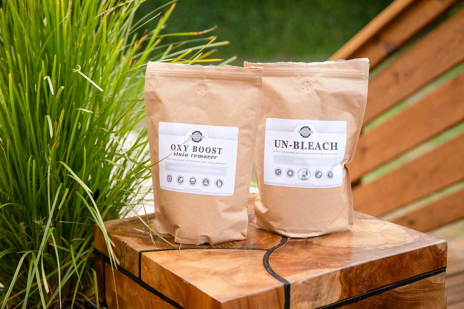 Two brown paper bags of cleaning products labeled 'Oxy Boost' and 'Un-Bleach' on a wooden table with a green plant on the side and a wooden fence in the background.