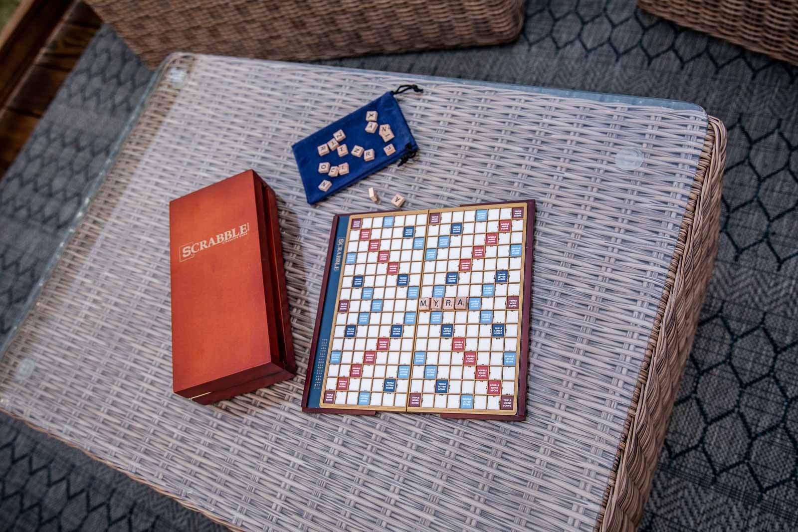 A Scrabble game set up with tiles spelling 'MYRA' on the board, a box of Scrabble tiles nearby, and a blue drawstring bag with unused tiles on a wicker table.