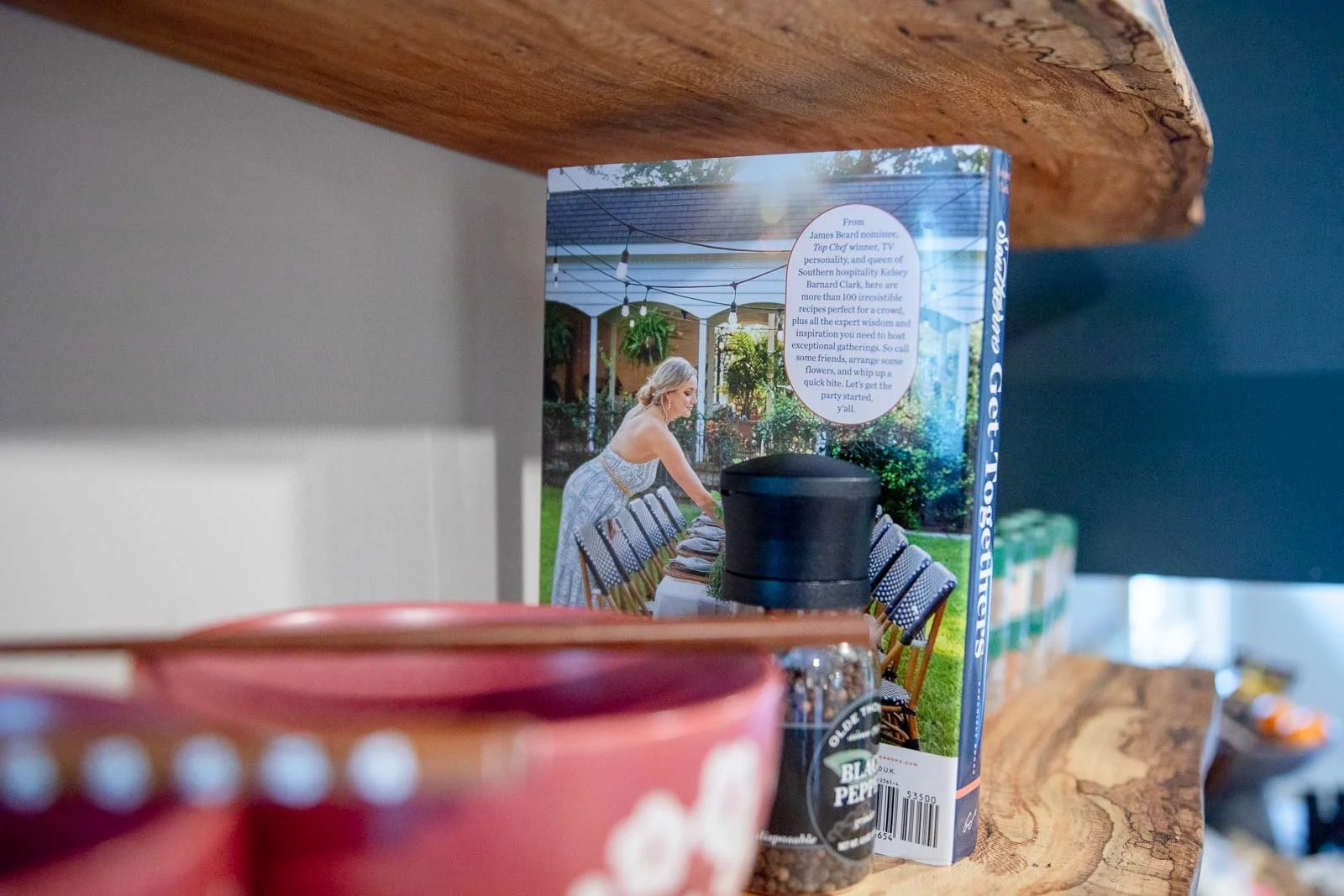 A magazine on a wooden shelf features a woman setting a long dining table outdoors with chairs and hanging plants, with a circular text overlay about a chef and hosting gatherings.