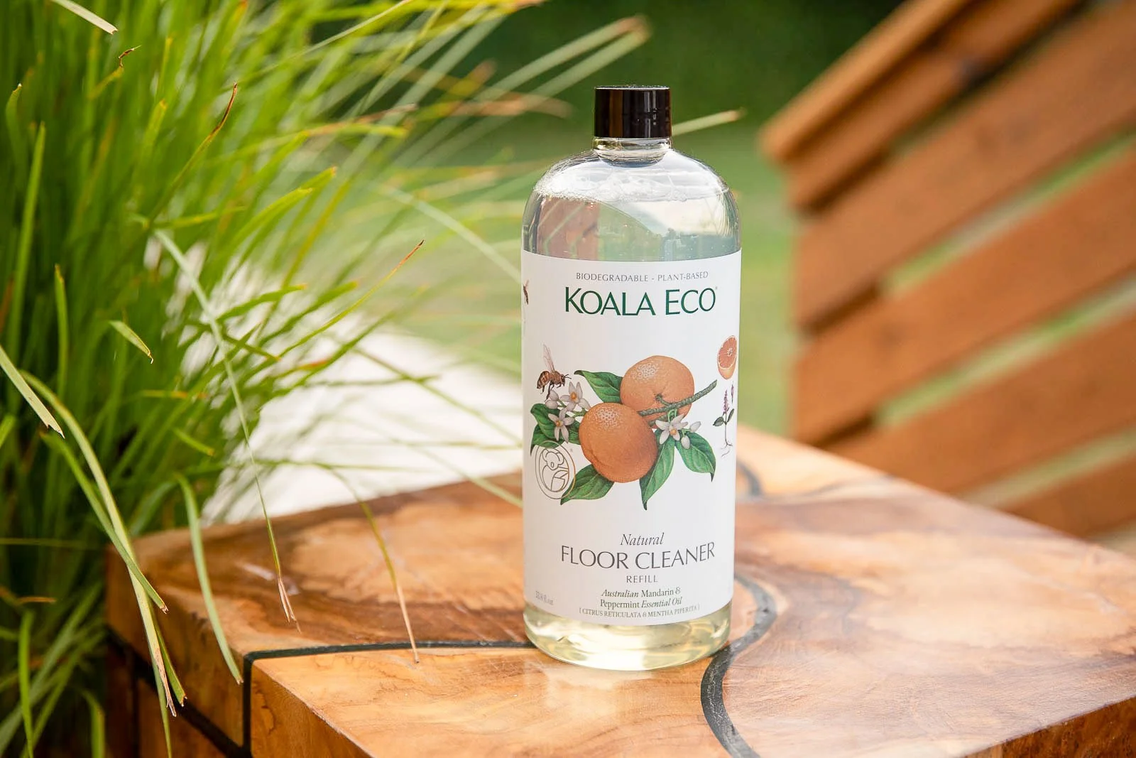 A clear plastic bottle of Koala Eco natural floor cleaner with a white label, illustrated with oranges, a bee, and white flowers, placed on a wooden surface outdoors with green plants and a brick background.