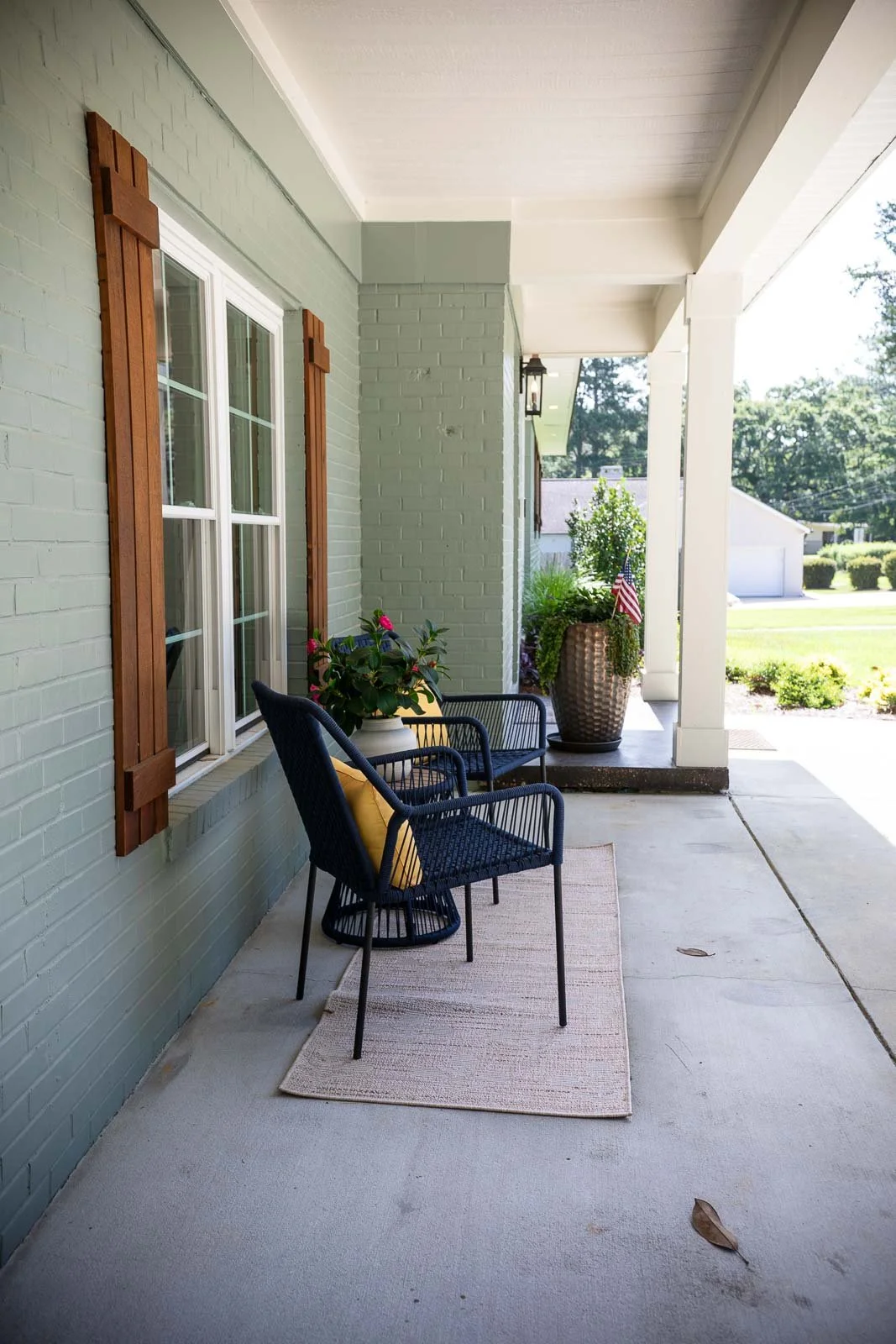 Front porch with black chairs, yellow pillows, potted plants, and an American flag in a large clay pot, overlooking a green lawn and house.